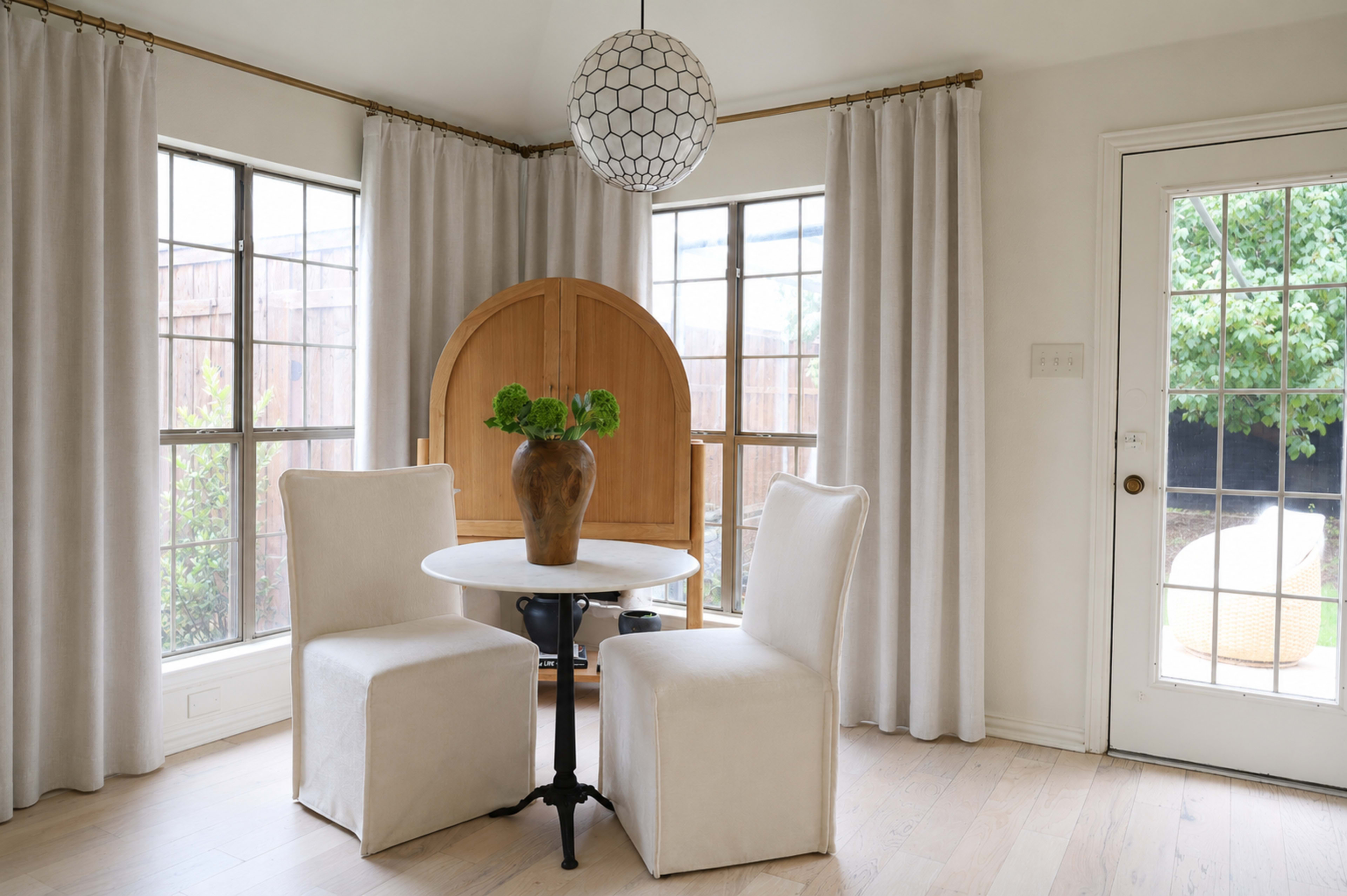 A round table with two upholstered chairs sits in a well-lit room, surrounded by large windows with sheer curtains and a wooden cabinet in the background.