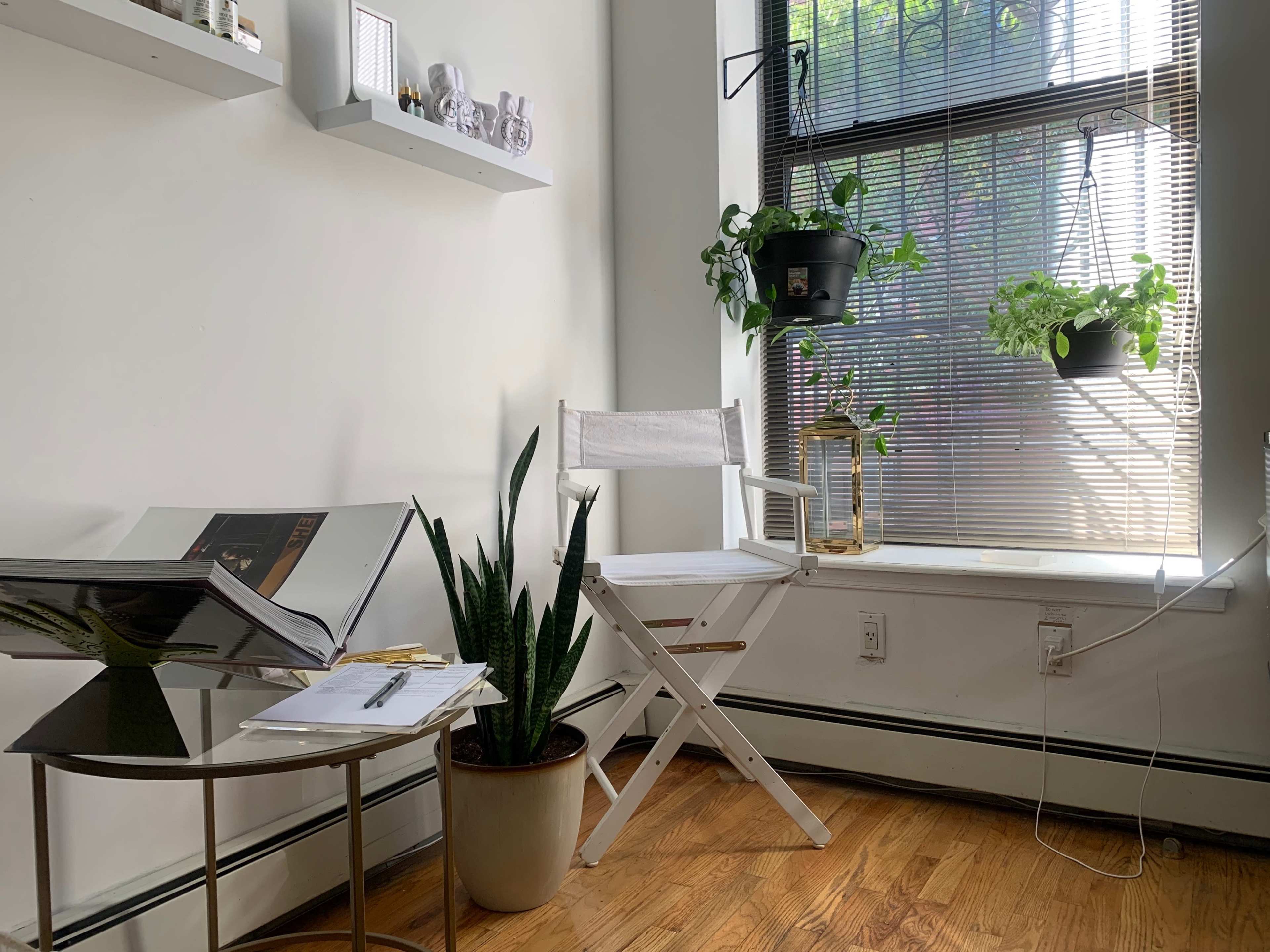 A white director's chair and a small side table are positioned beside a window with potted plants, in a sunlit corner of a room.
