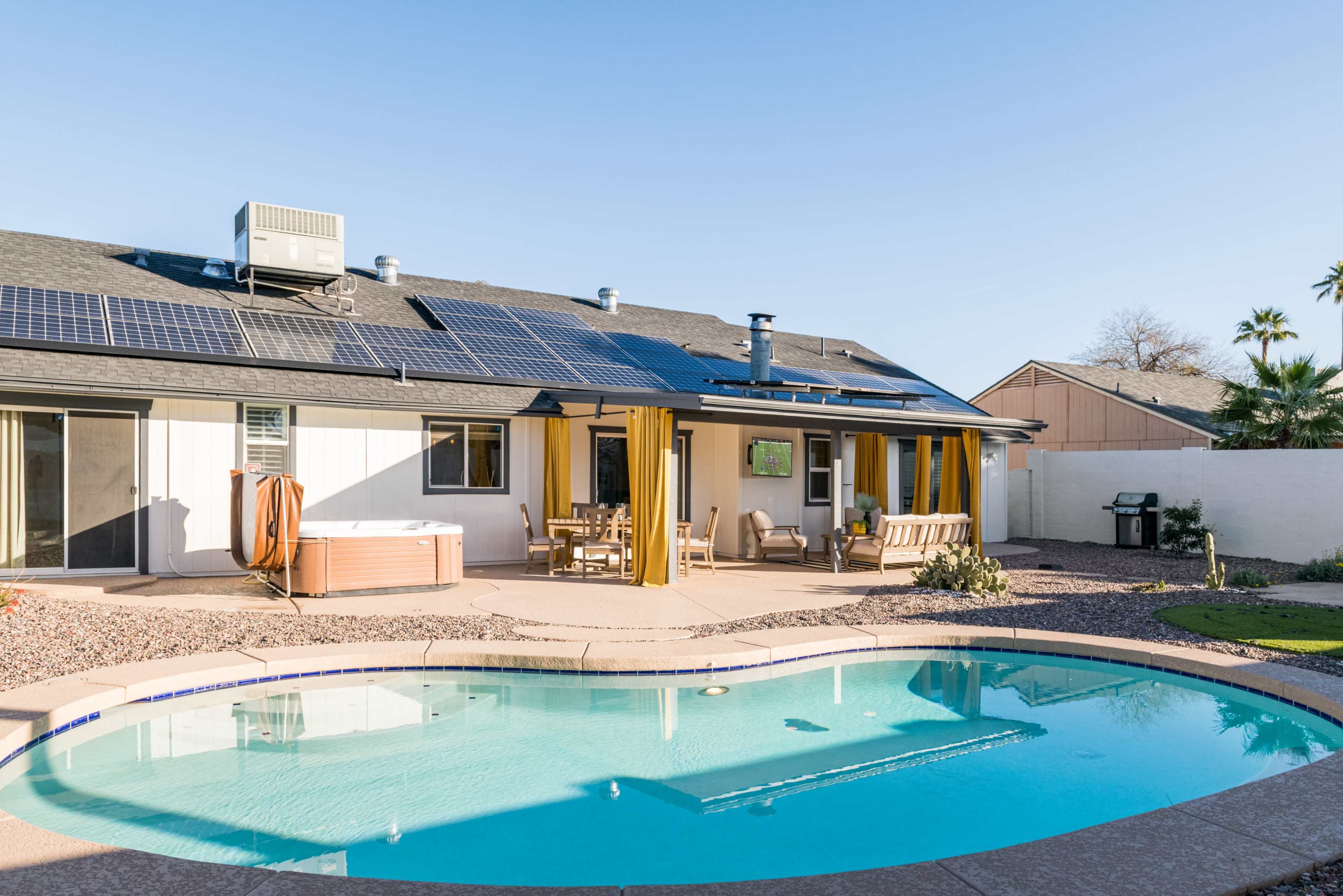 A backyard features a swimming pool, hot tub, and a house with solar panels and yellow curtains.