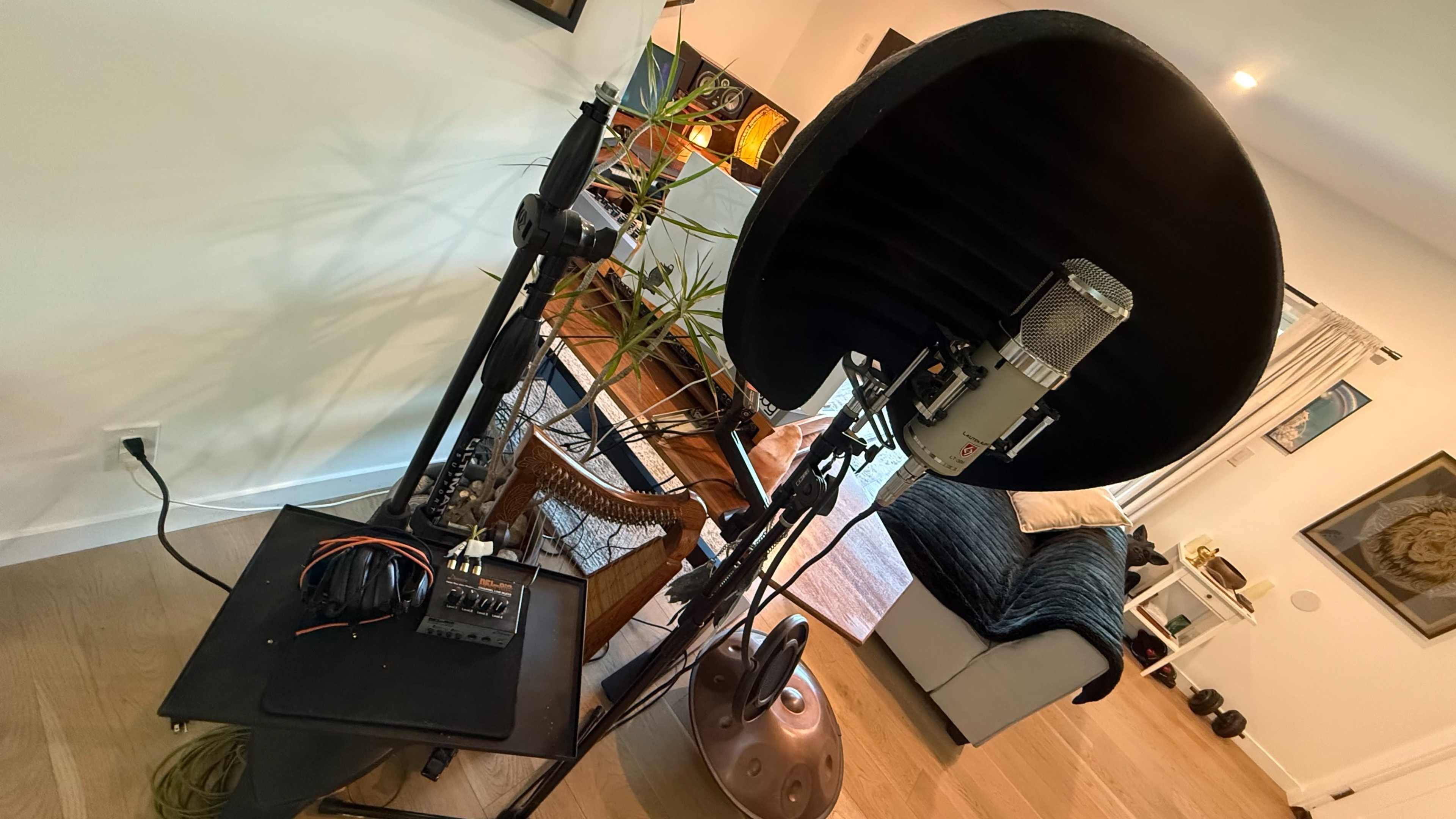 A large microphone with a reflection shield is positioned on a stand next to a table with audio equipment in a well-lit room.
