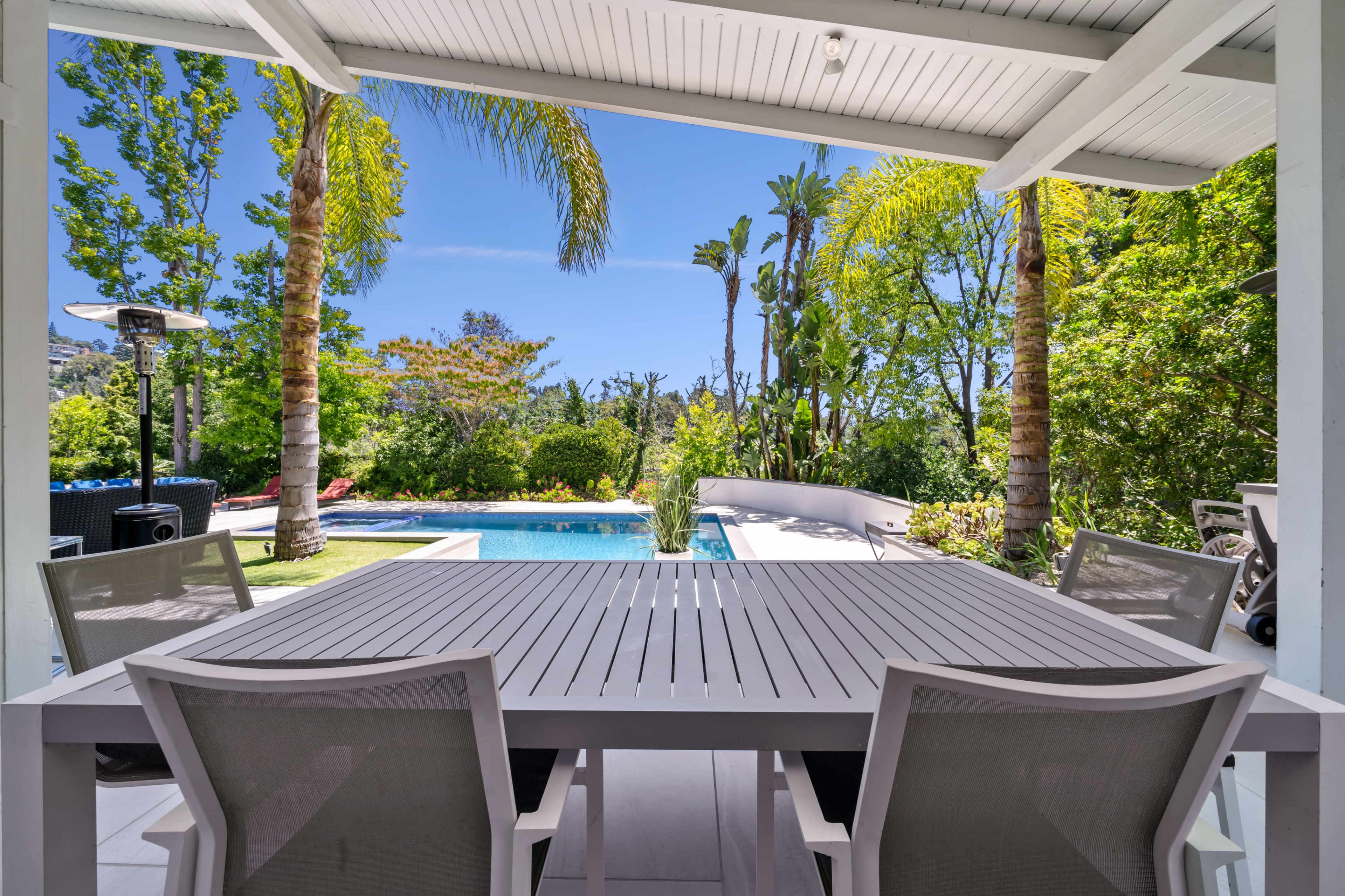 The image shows a patio with a white outdoor dining table and chairs, overlooking a landscaped garden and swimming pool.