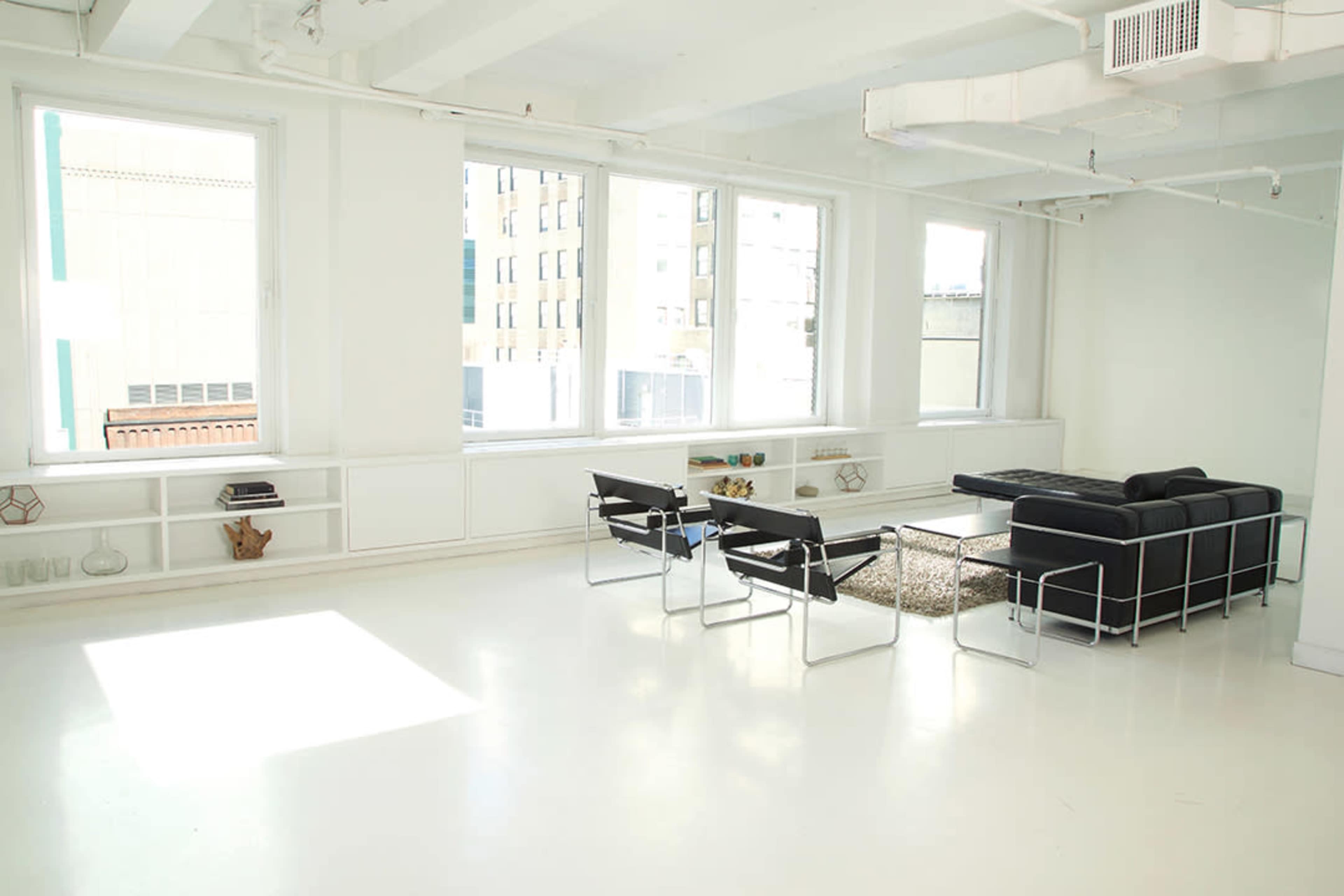 A bright, minimalistic room with large windows, a black sofa, several black chairs, and a low coffee table placed on a patterned rug.