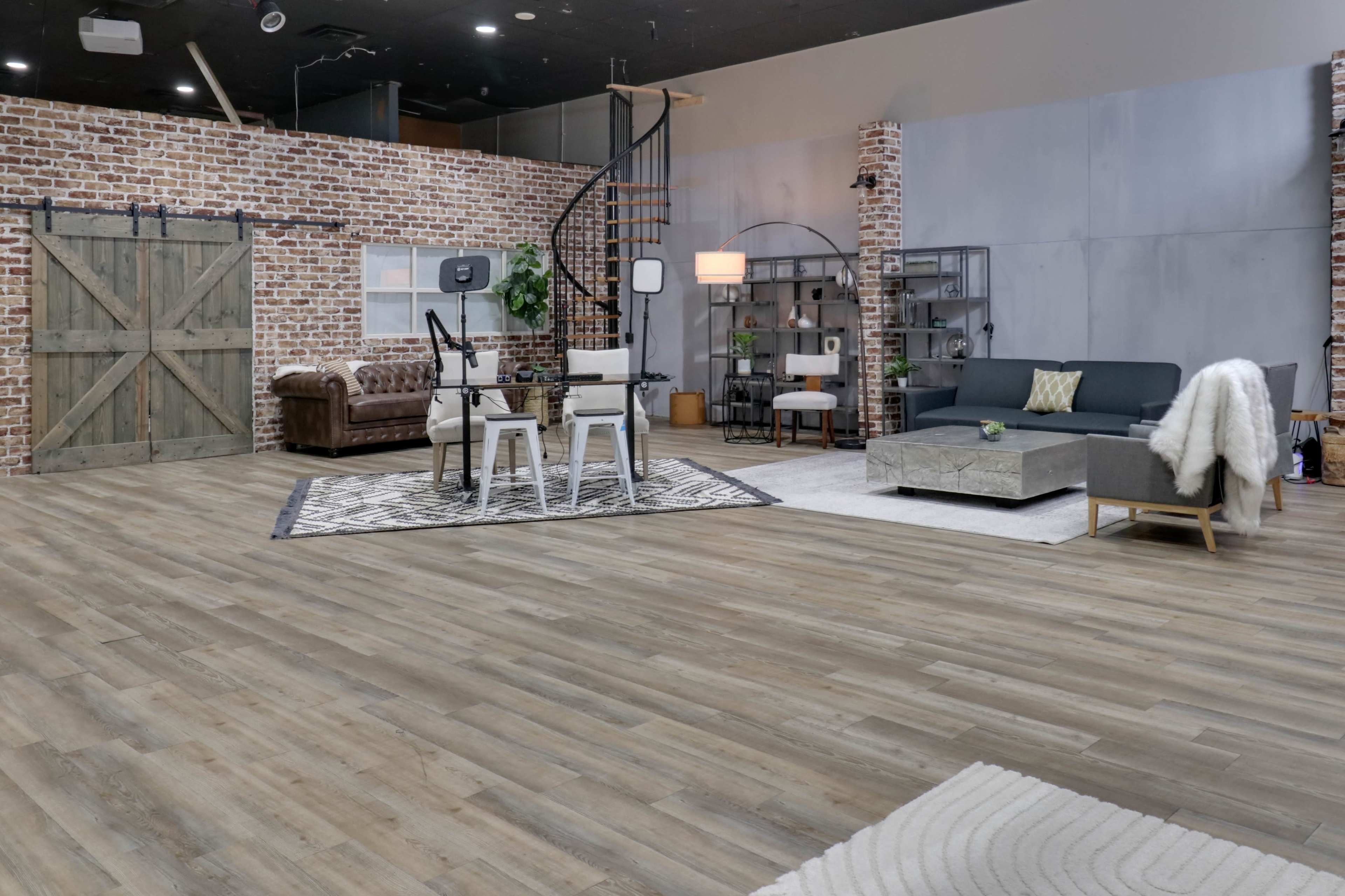The image shows a spacious interior living area with a mix of modern and rustic decor, featuring a brown couch, a gray sectional, a patterned rug, and brick walls.
