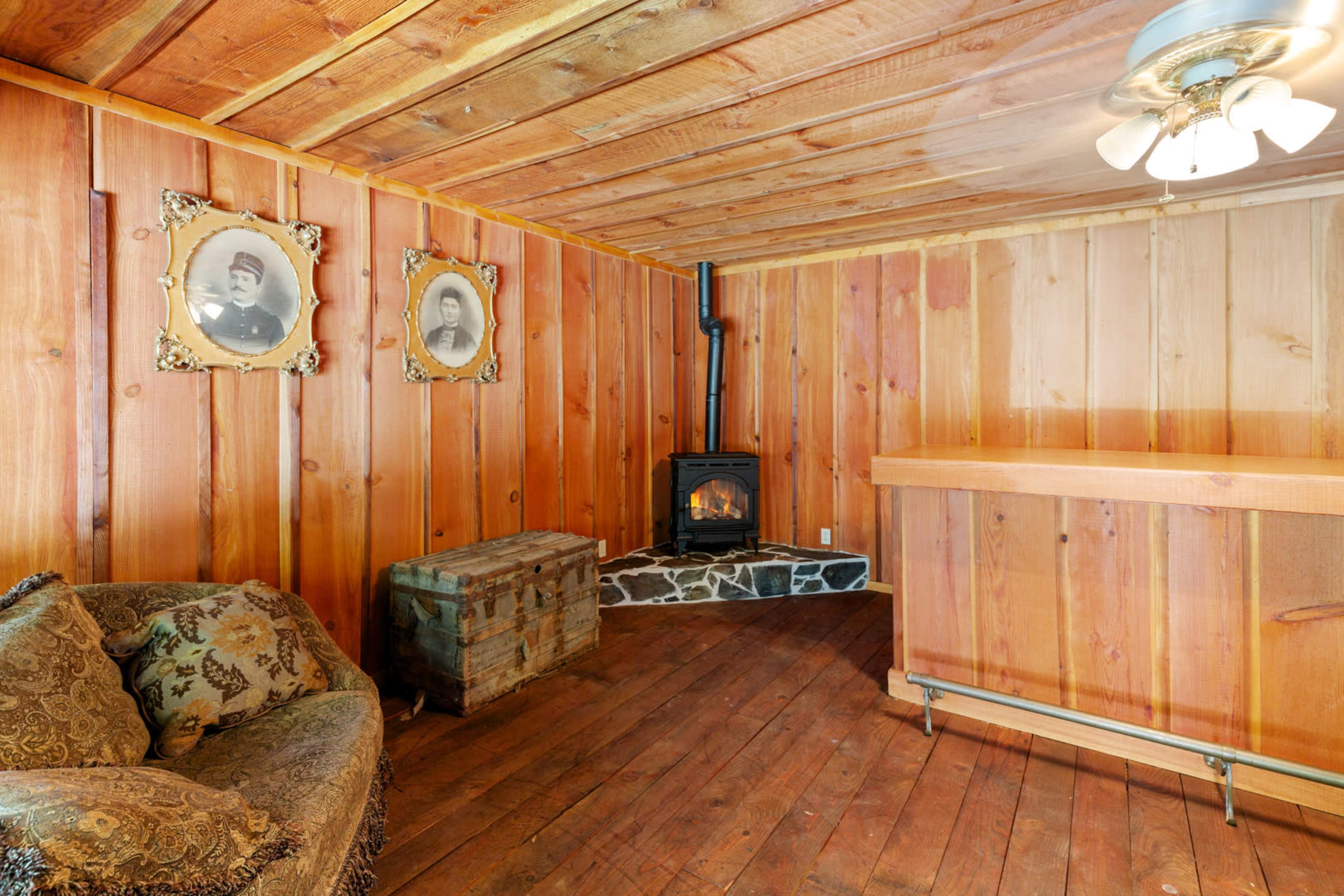 The image shows a rustic wooden room with a small stove, a sofa, a wooden chest, and two framed portraits on the wall.