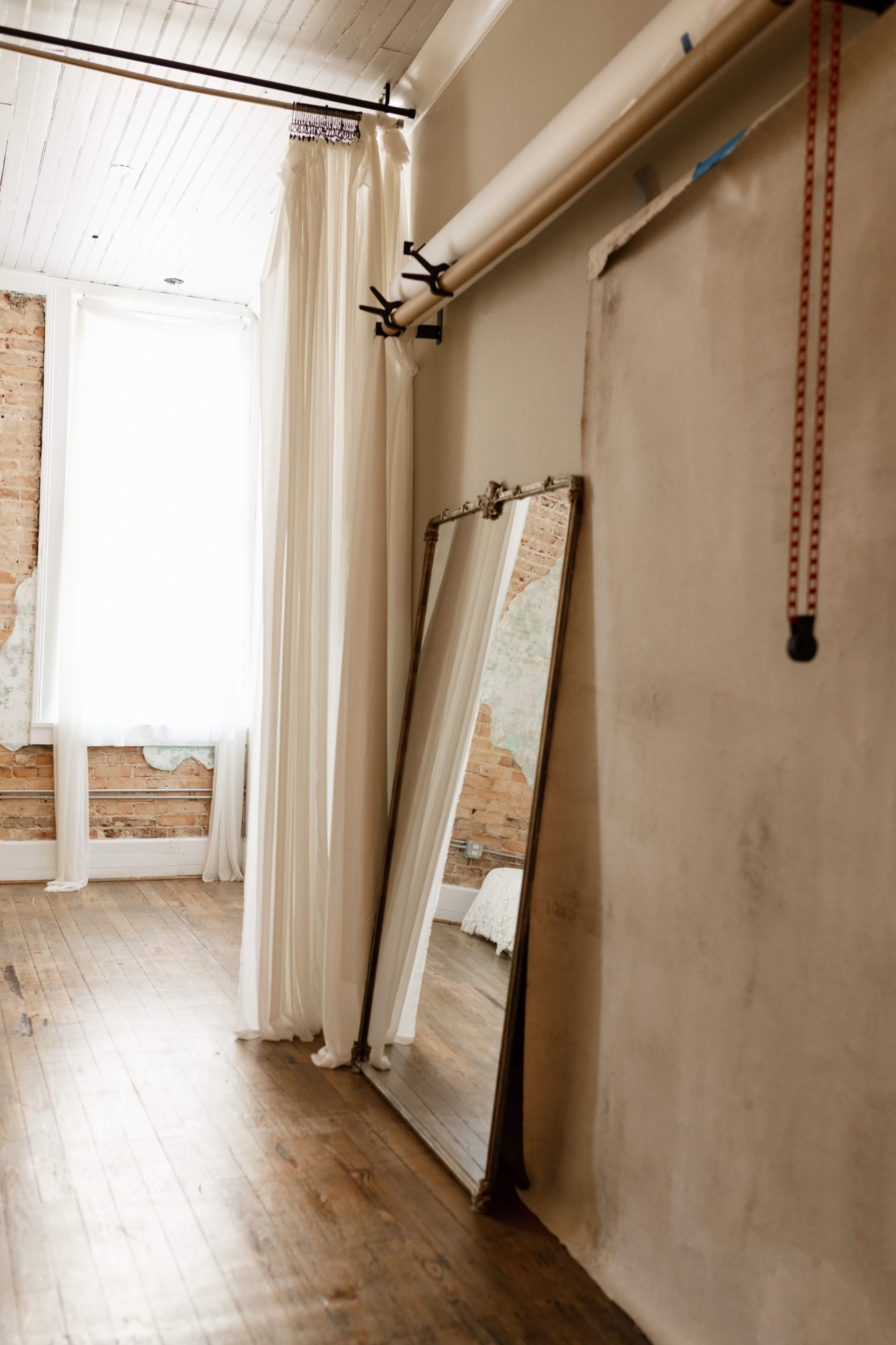 The image features a minimalist room with wooden floors, a large mirror against a wall, sheer curtains near a window, and a backdrop hanging from a pipe.