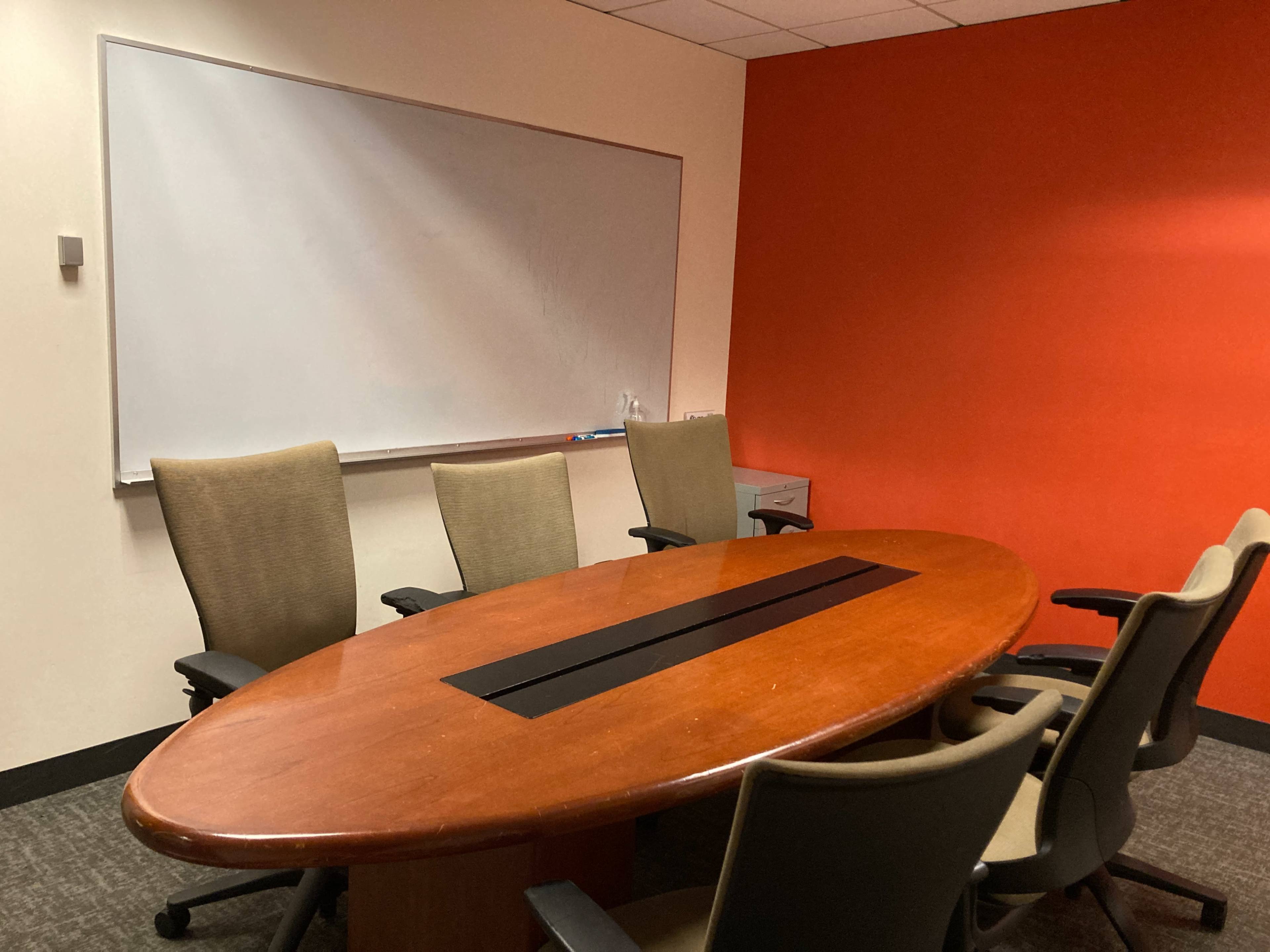 A conference room features a large oval wooden table surrounded by six chairs, with a whiteboard on one wall and an orange accent wall.