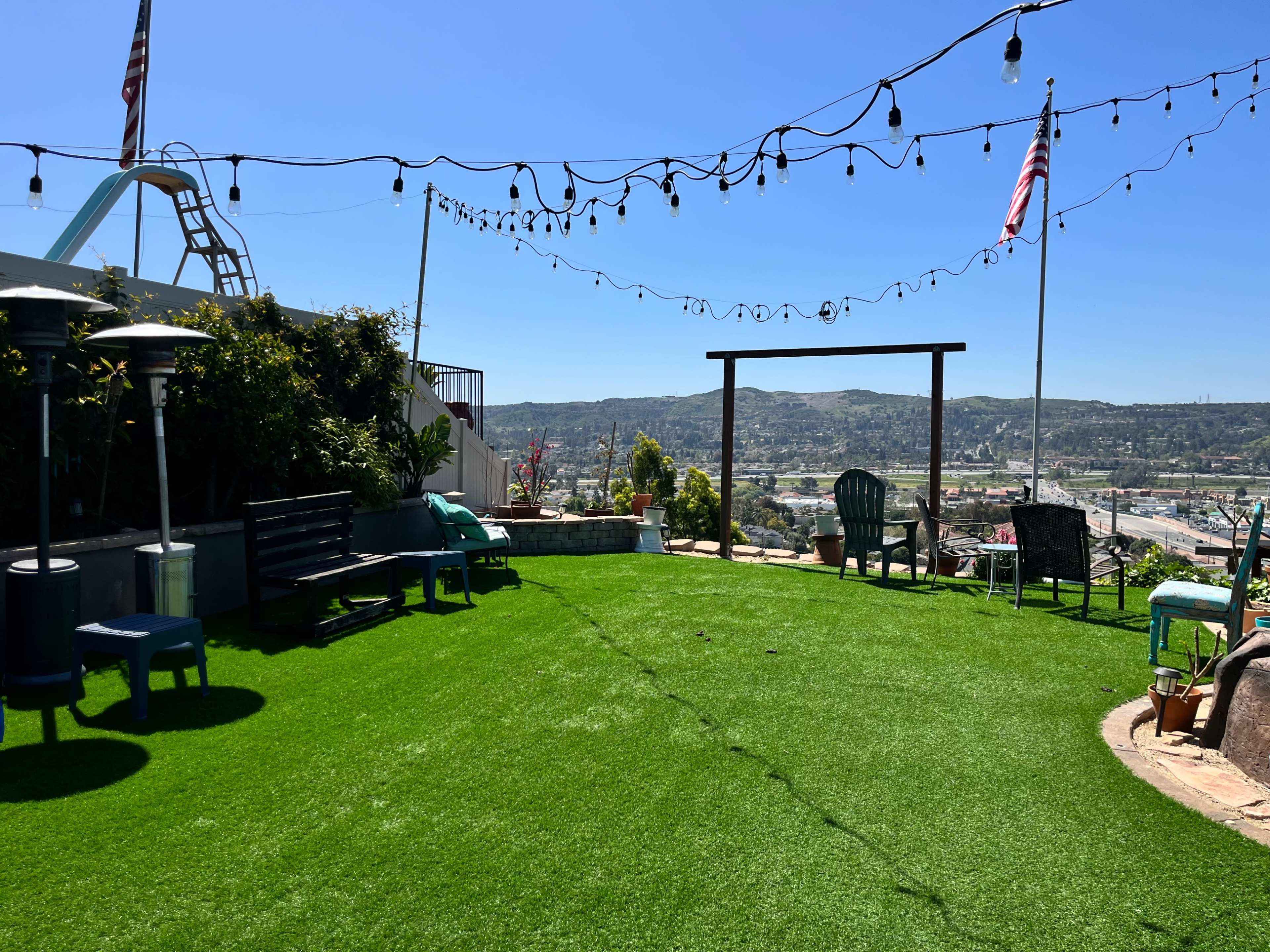 The image shows a landscaped garden area with artificial turf, outdoor seating, and string lights above, overlooking a scenic view.