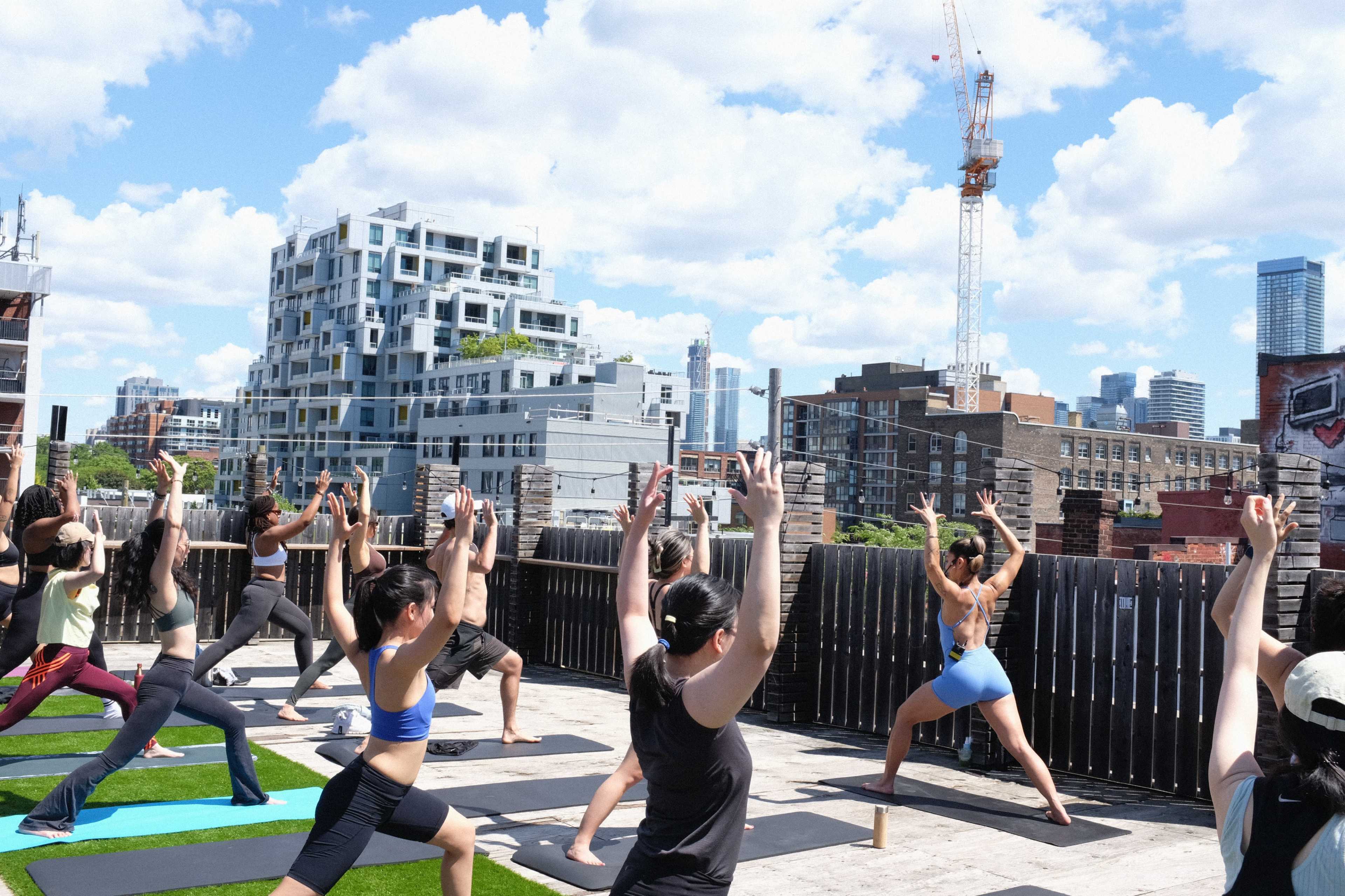 A group of individuals is performing yoga on a rooftop with a city skyline and construction crane in the background.