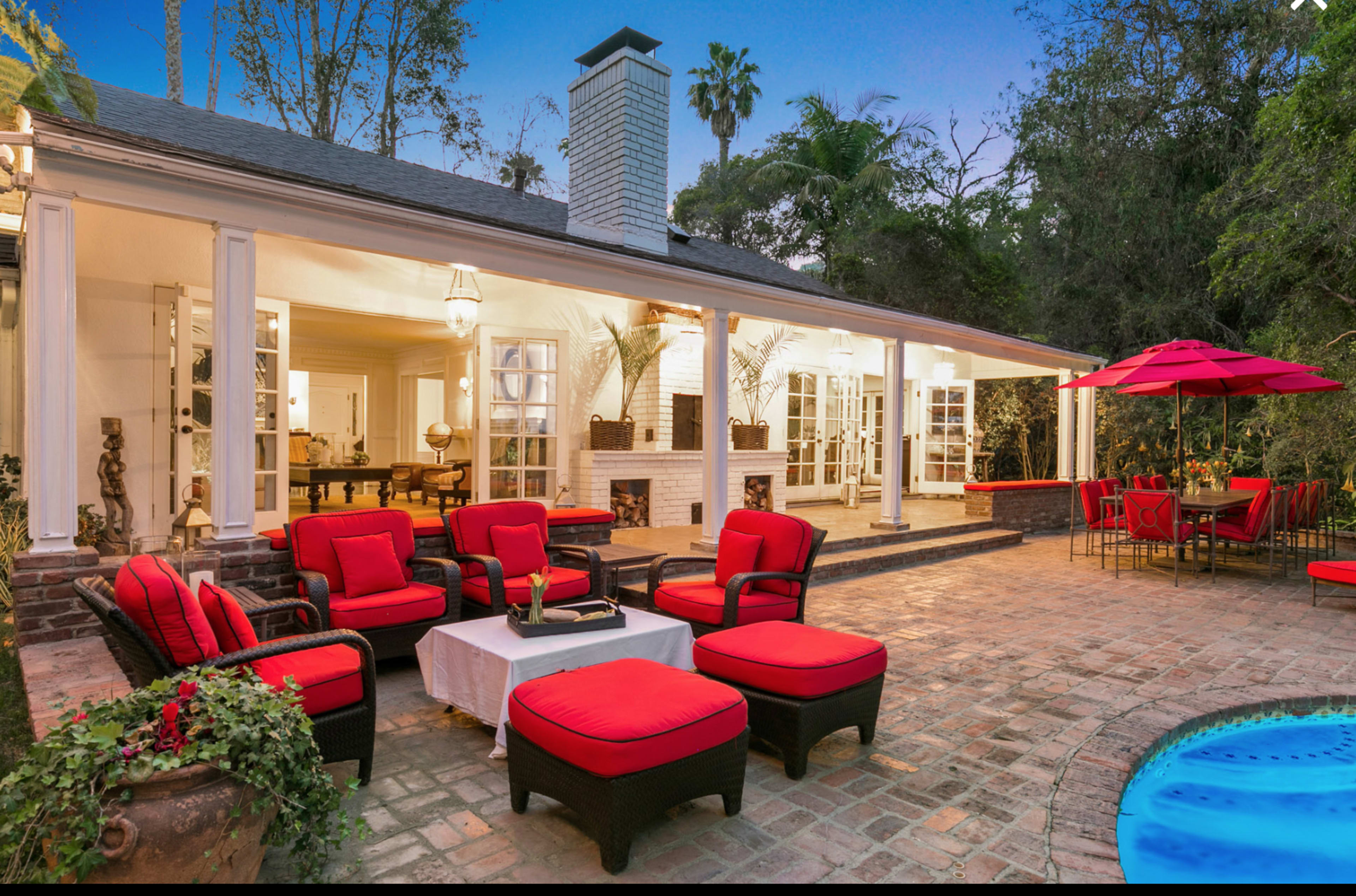 The image shows a cozy outdoor seating area with red cushions, adjacent to a pool and a dining space, all set in a lush, green environment.