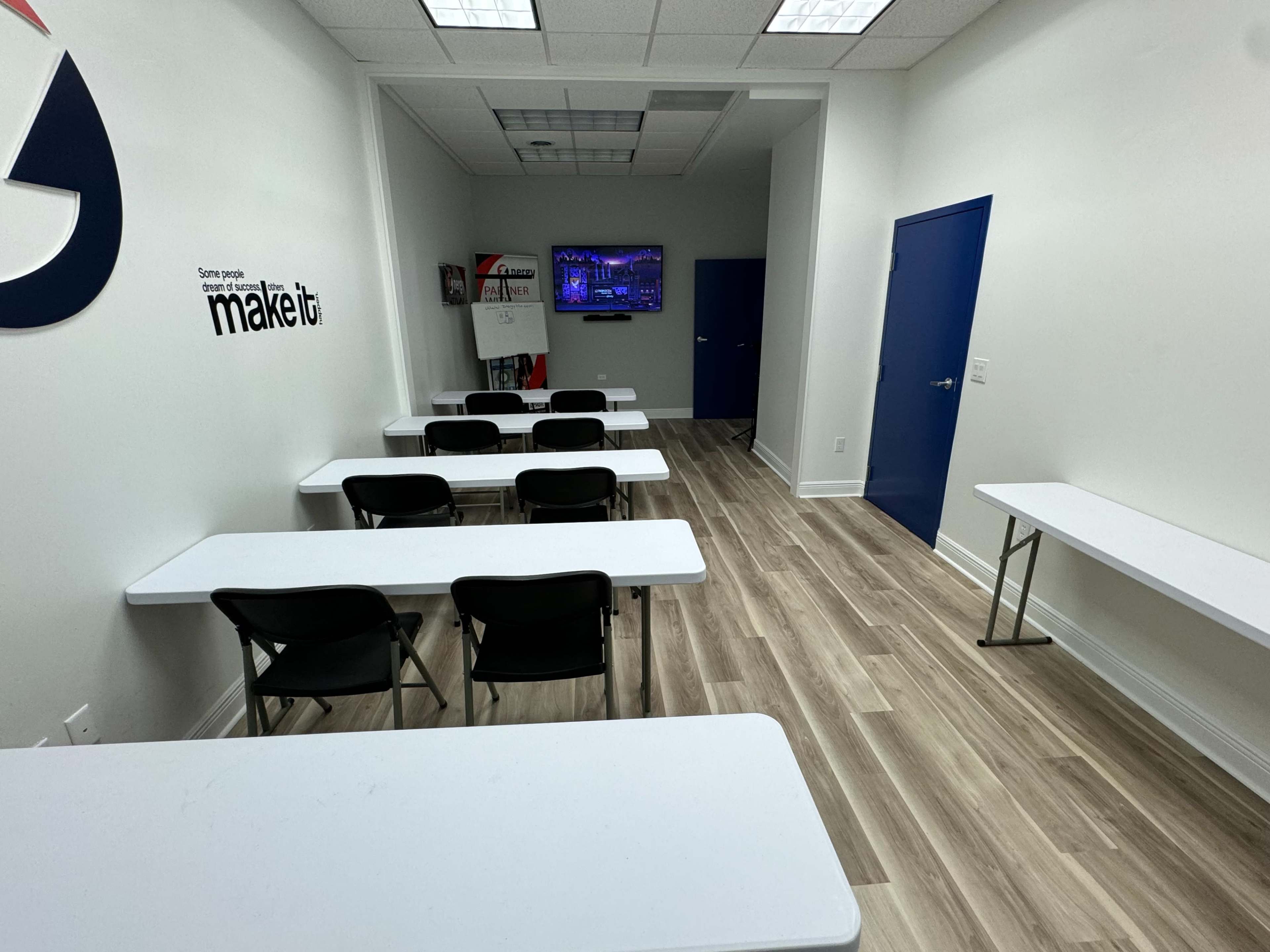The image shows a classroom setup with several white tables and chairs arranged neatly, a whiteboard, and a television mounted on the wall.