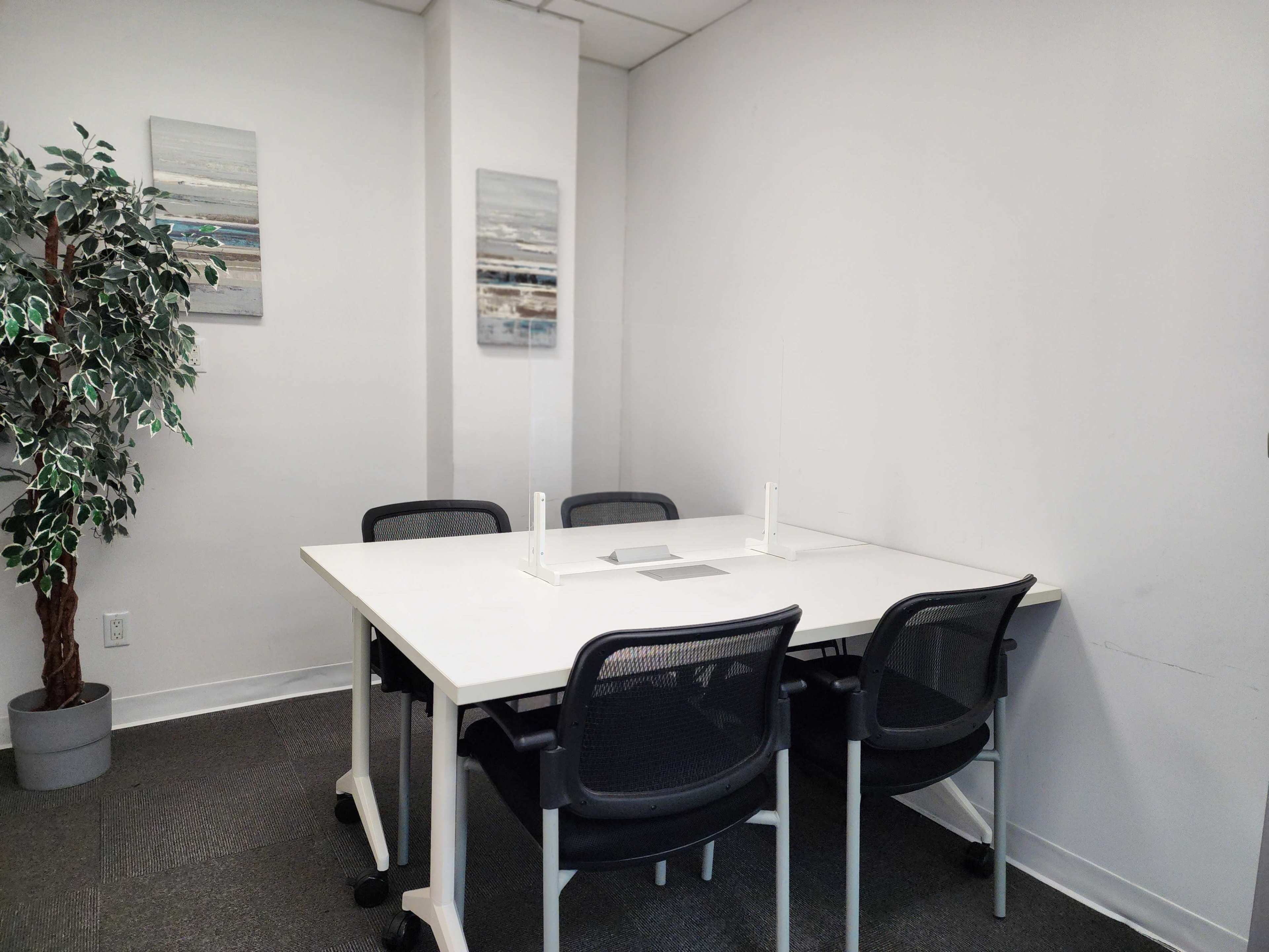 A small, minimalist meeting room featuring a white table surrounded by four black chairs, a potted plant, and two framed artworks on the wall.