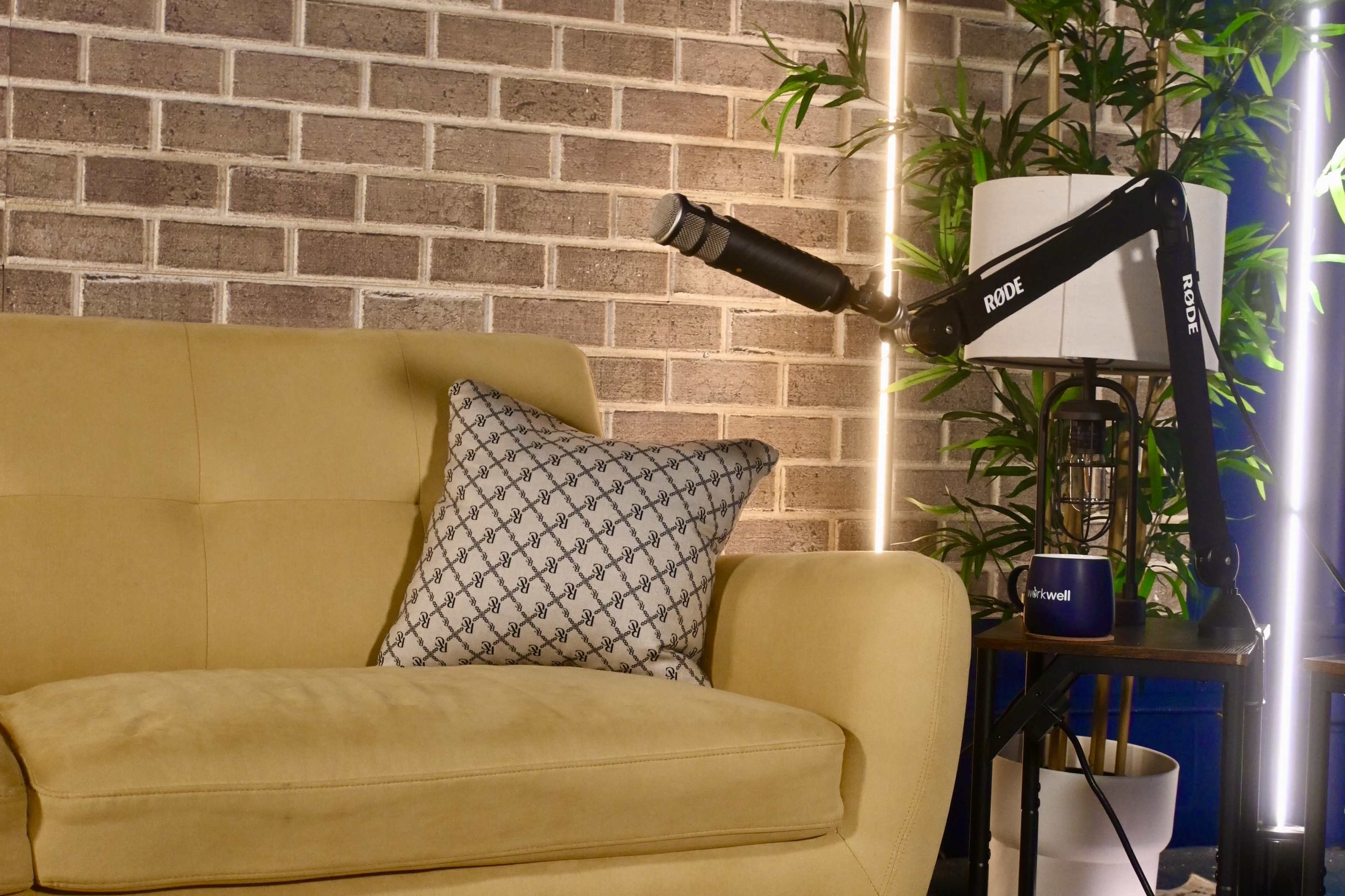 A yellow couch with a decorative pillow sits next to a side table featuring a microphone and a potted plant against a brick wall.