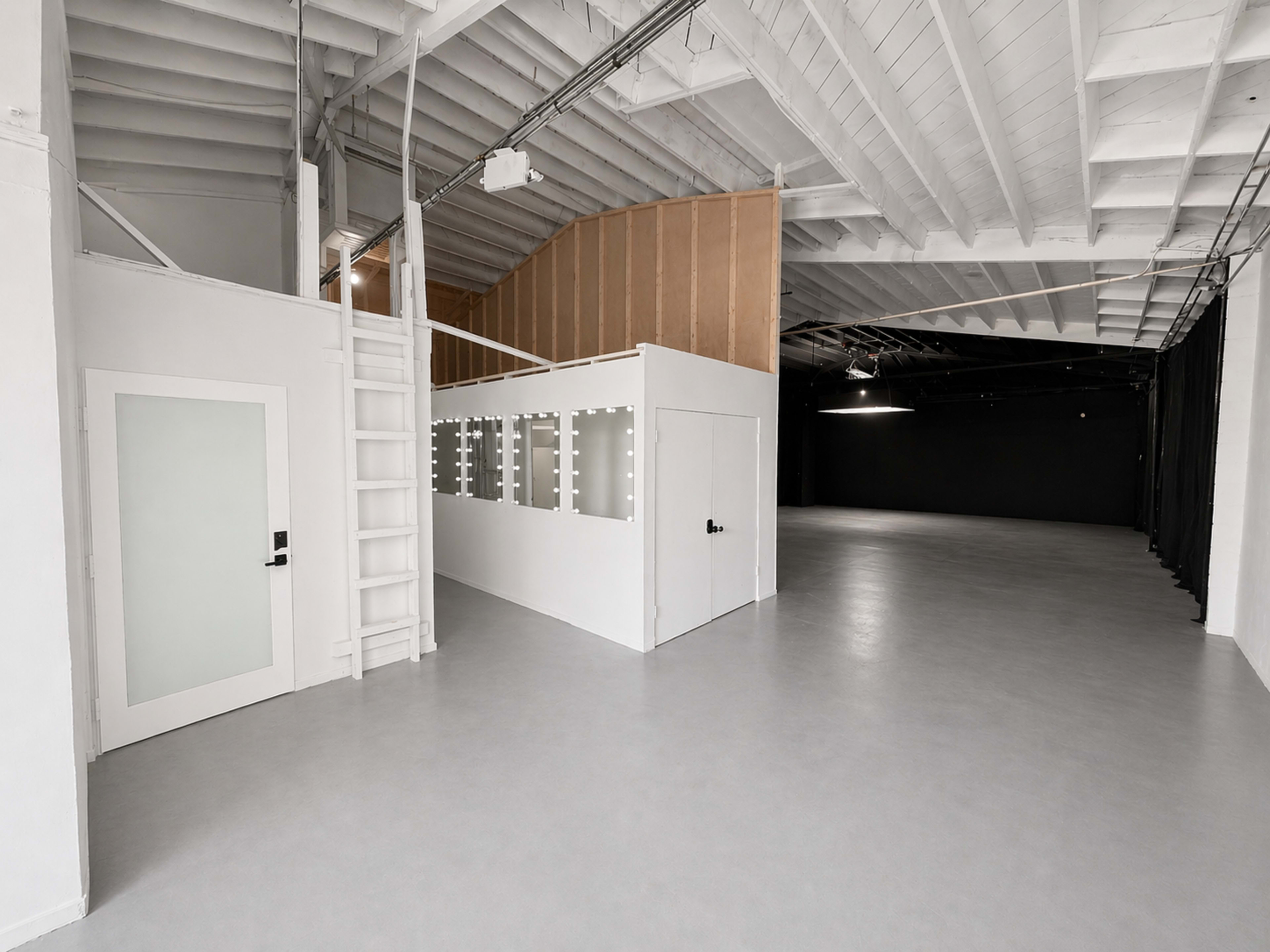 The image features a spacious, empty interior of a building with white walls, high ceilings, and a staircase leading to a small loft area.
