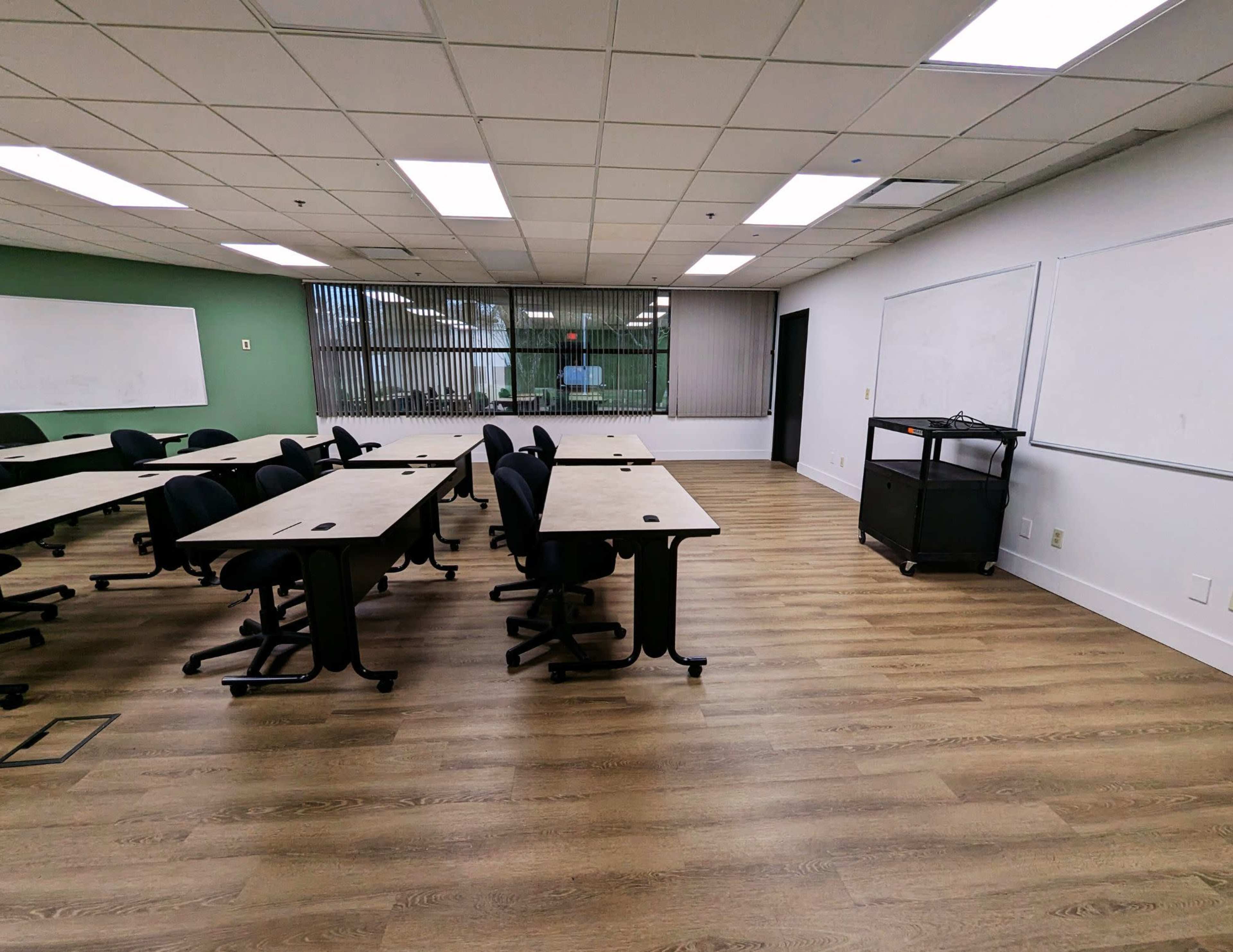 A classroom features several tables arranged with chairs, large windows overlooking greenery, and whiteboards on two walls.