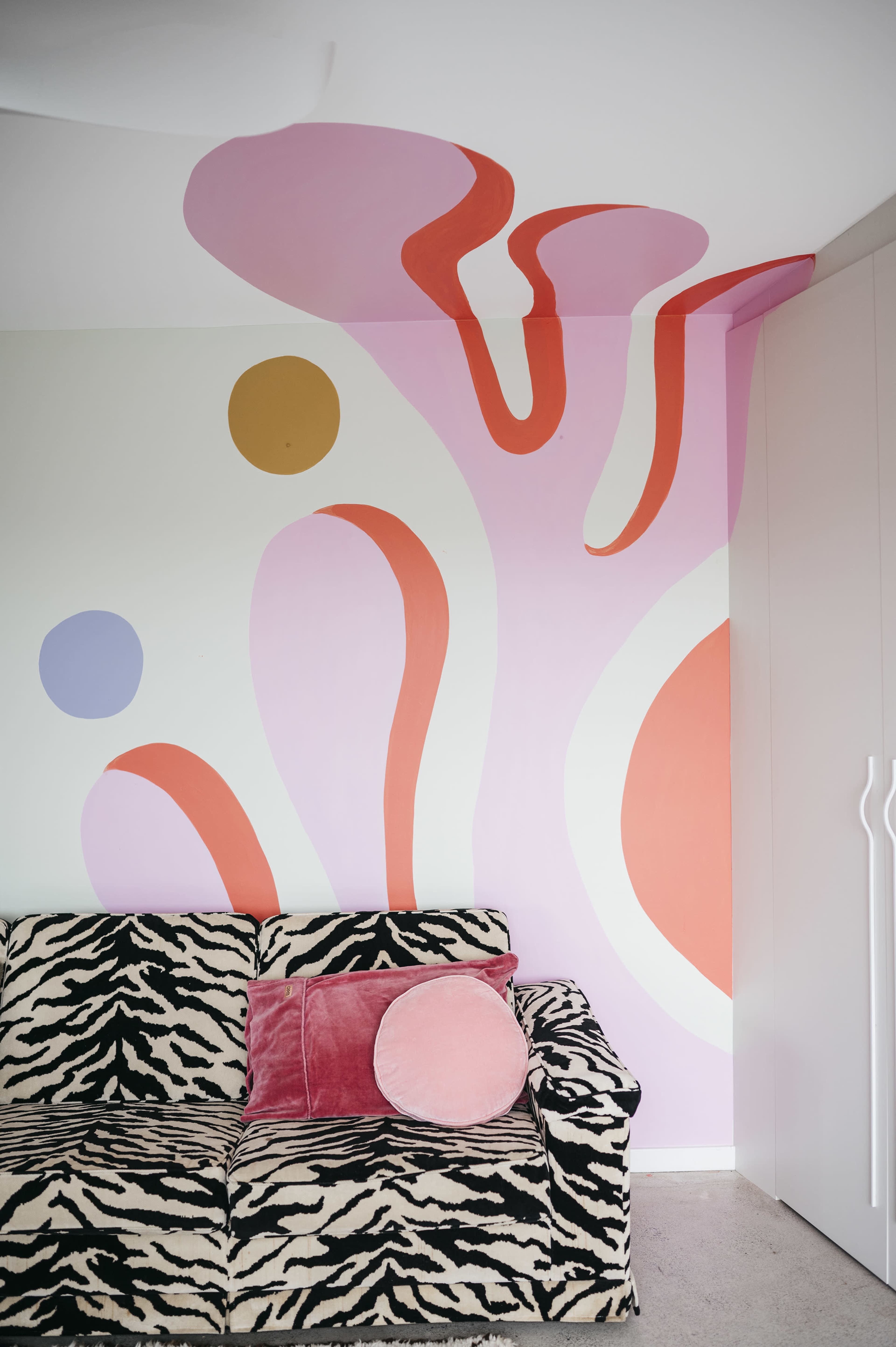 A zebra-striped sofa with a pink pillow against a wall featuring abstract pastel-colored shapes and patterns.