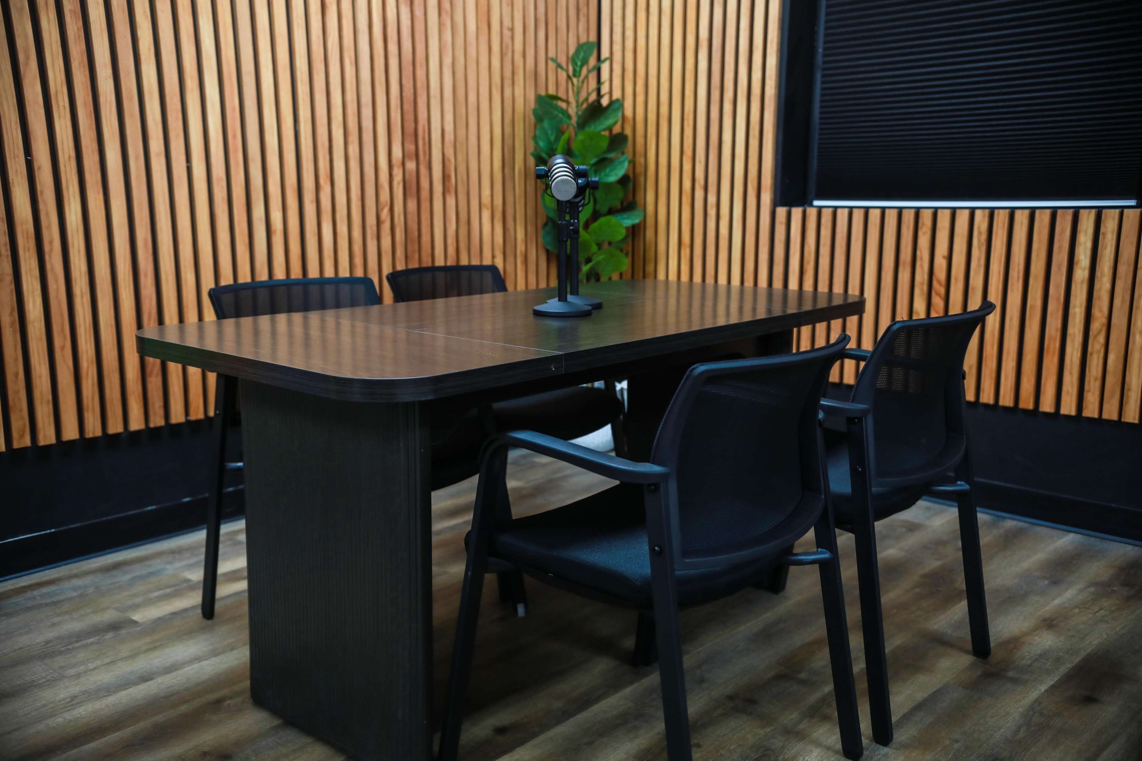 A conference room features a dark wooden table with a microphone on it, surrounded by black chairs against a backdrop of vertical wooden slats.