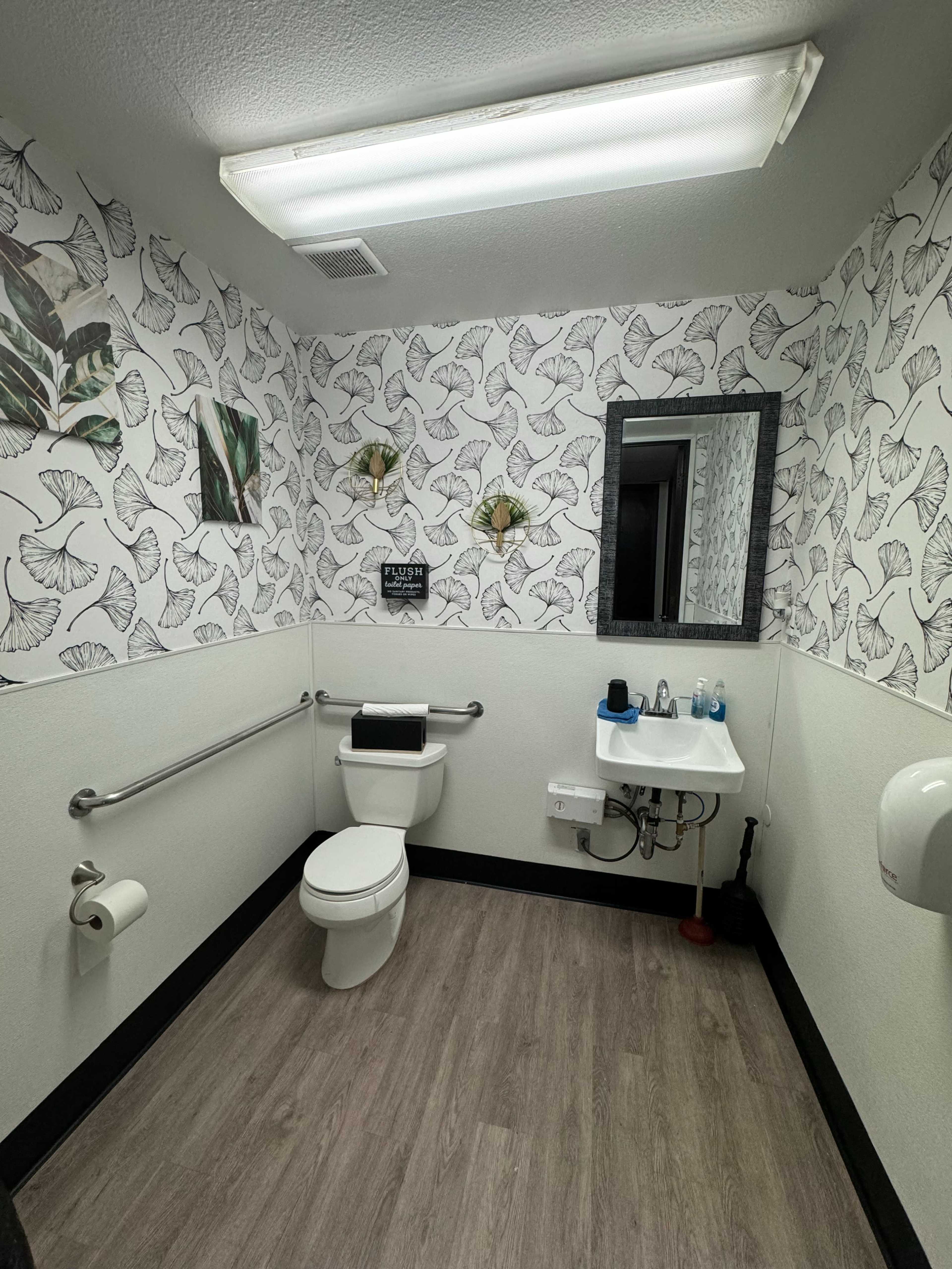 A restroom with decorative leaf-patterned wallpaper, a toilet, a sink with a mirror, and safety grab bars on the walls.