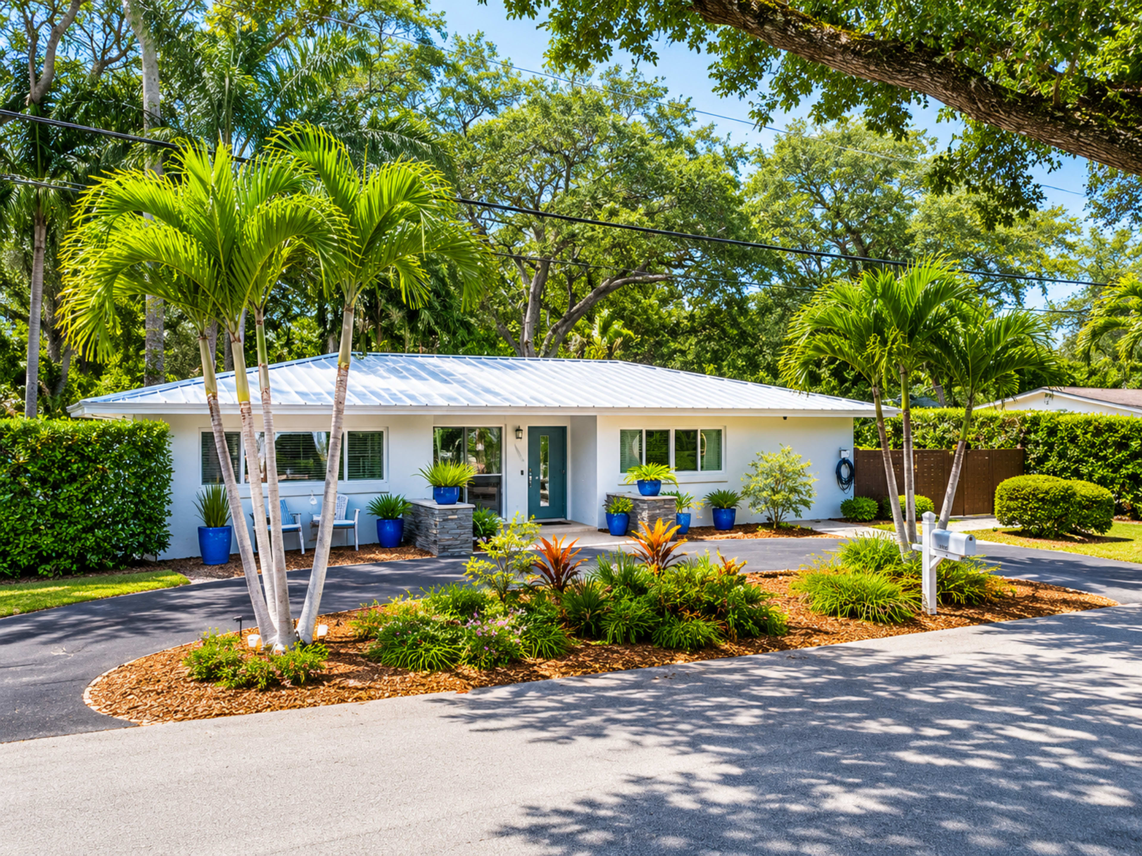 A single-story house with a light blue exterior features a landscaped front yard with palm trees and colorful plants, bordered by a driveway.