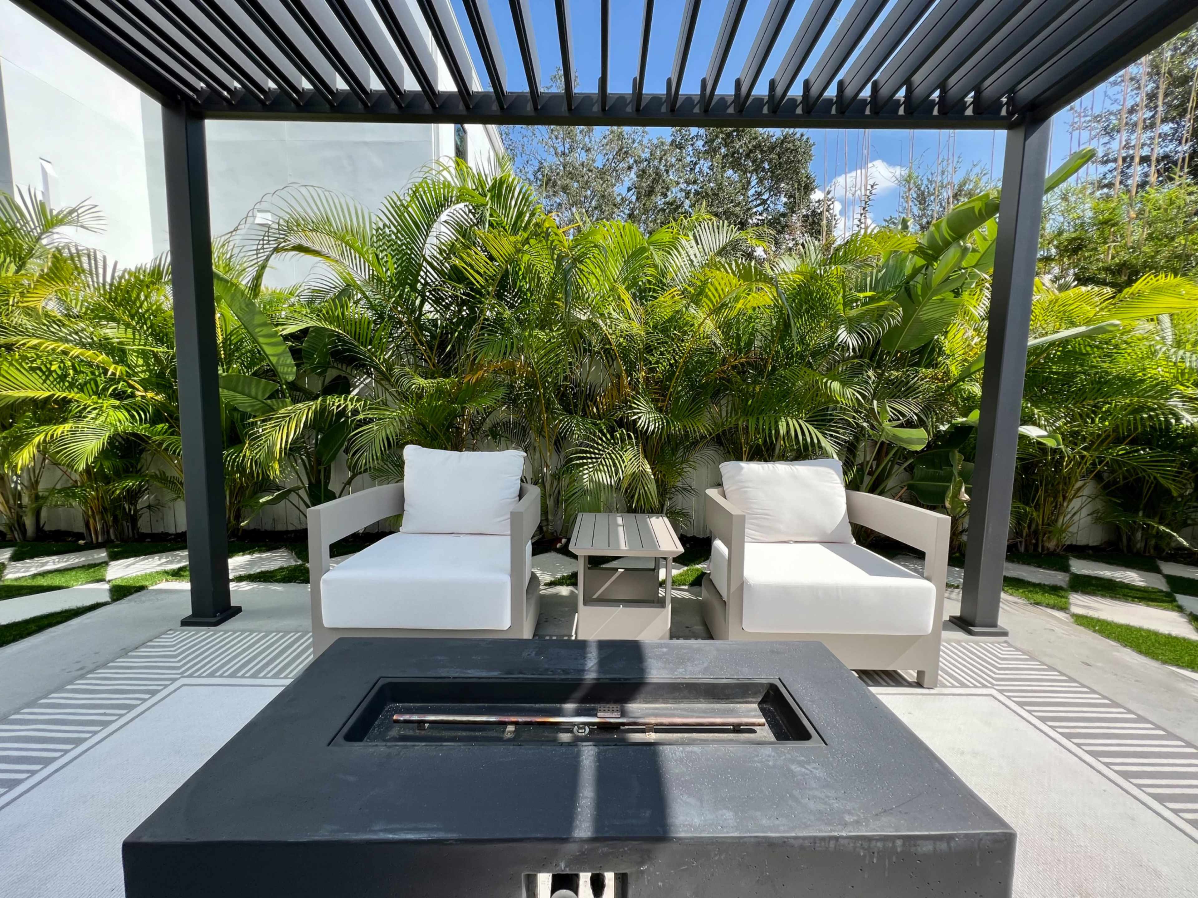 A cozy outdoor seating area features two white chairs and a small table under a pergola, surrounded by lush greenery.