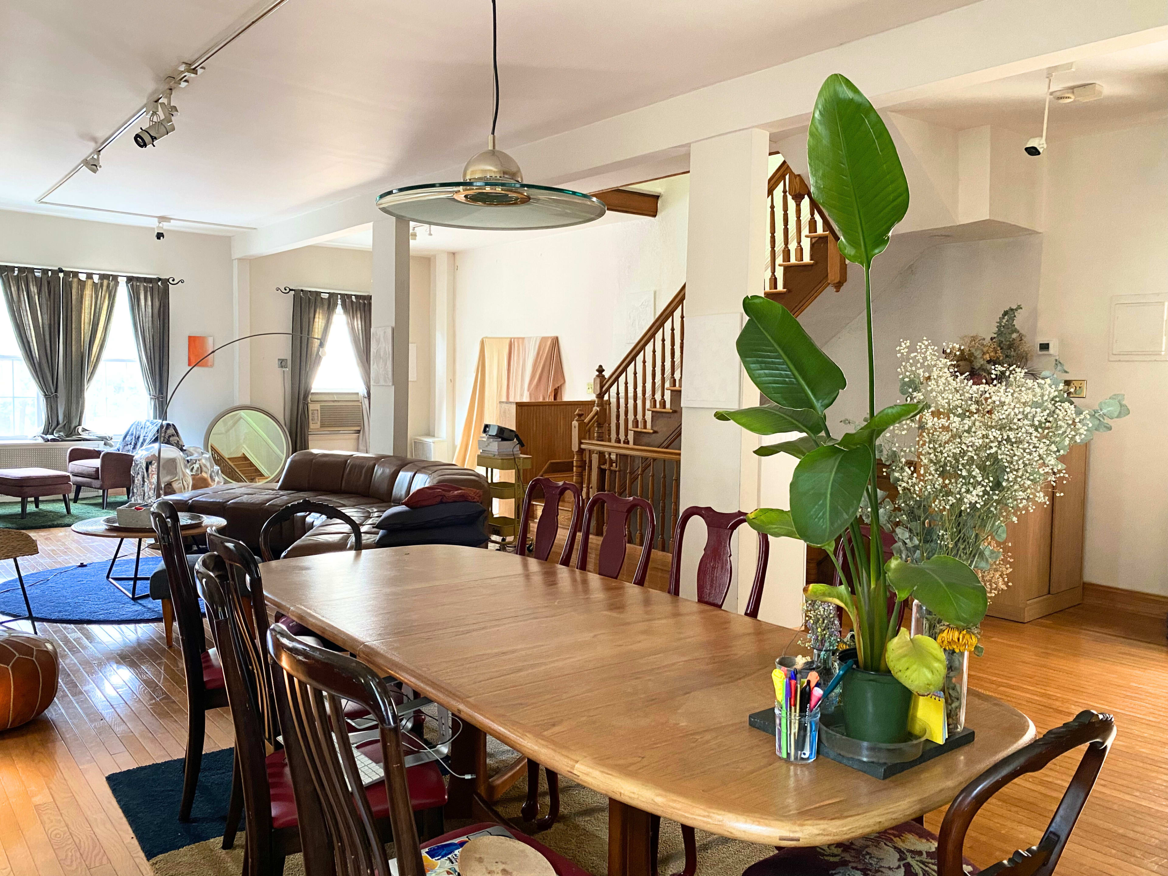A spacious dining area with a long wooden table, surrounded by chairs, and connected to a living space featuring a sofa and a staircase in the background.