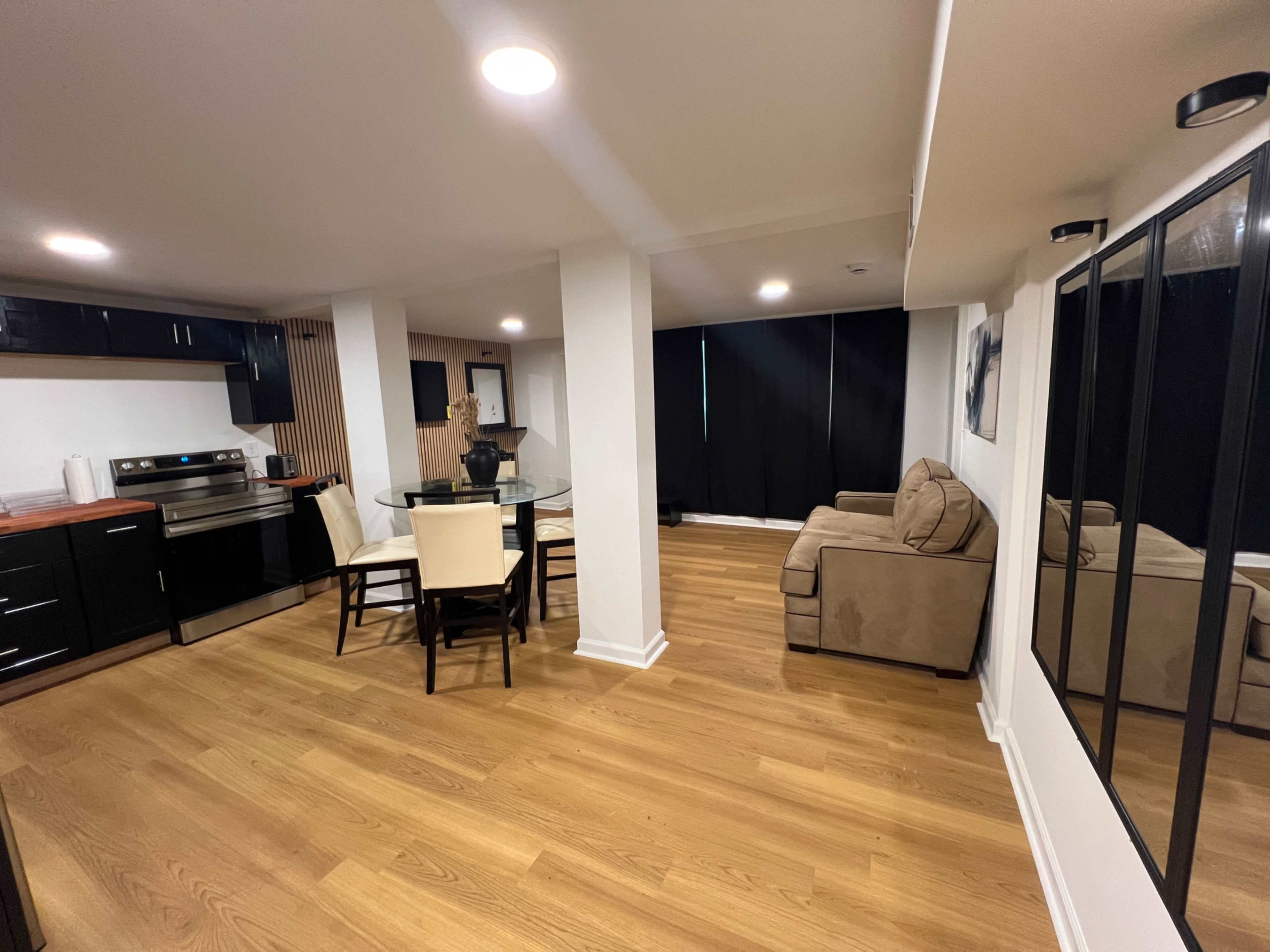 The image shows a modern kitchen and living area with hardwood floors, a circular dining table with six chairs, a beige couch, and large black curtains covering the windows.