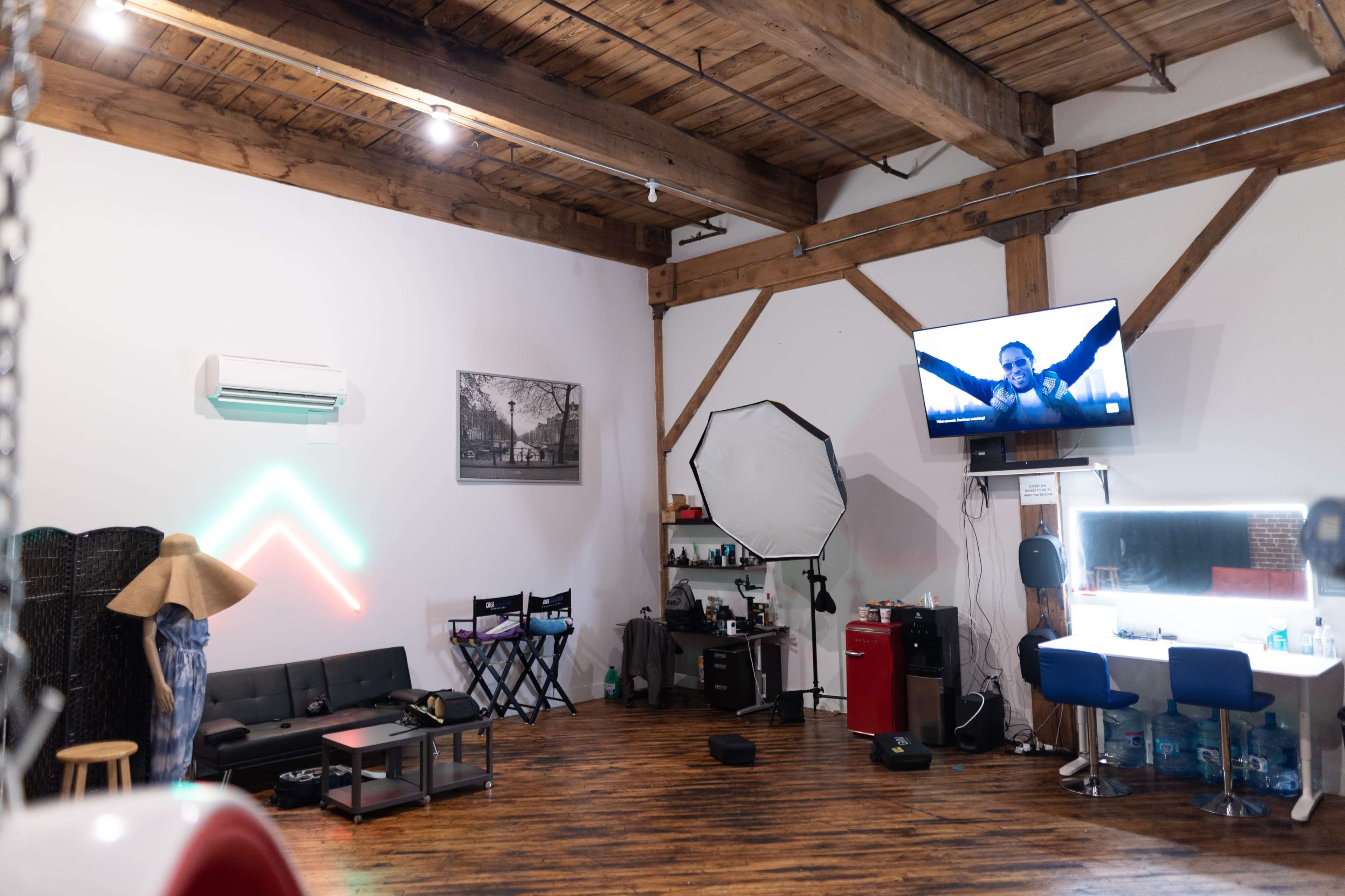 A spacious studio with wooden beams, a flat-screen TV displaying a person, a black couch, a makeup station, and various equipment organized along the walls.