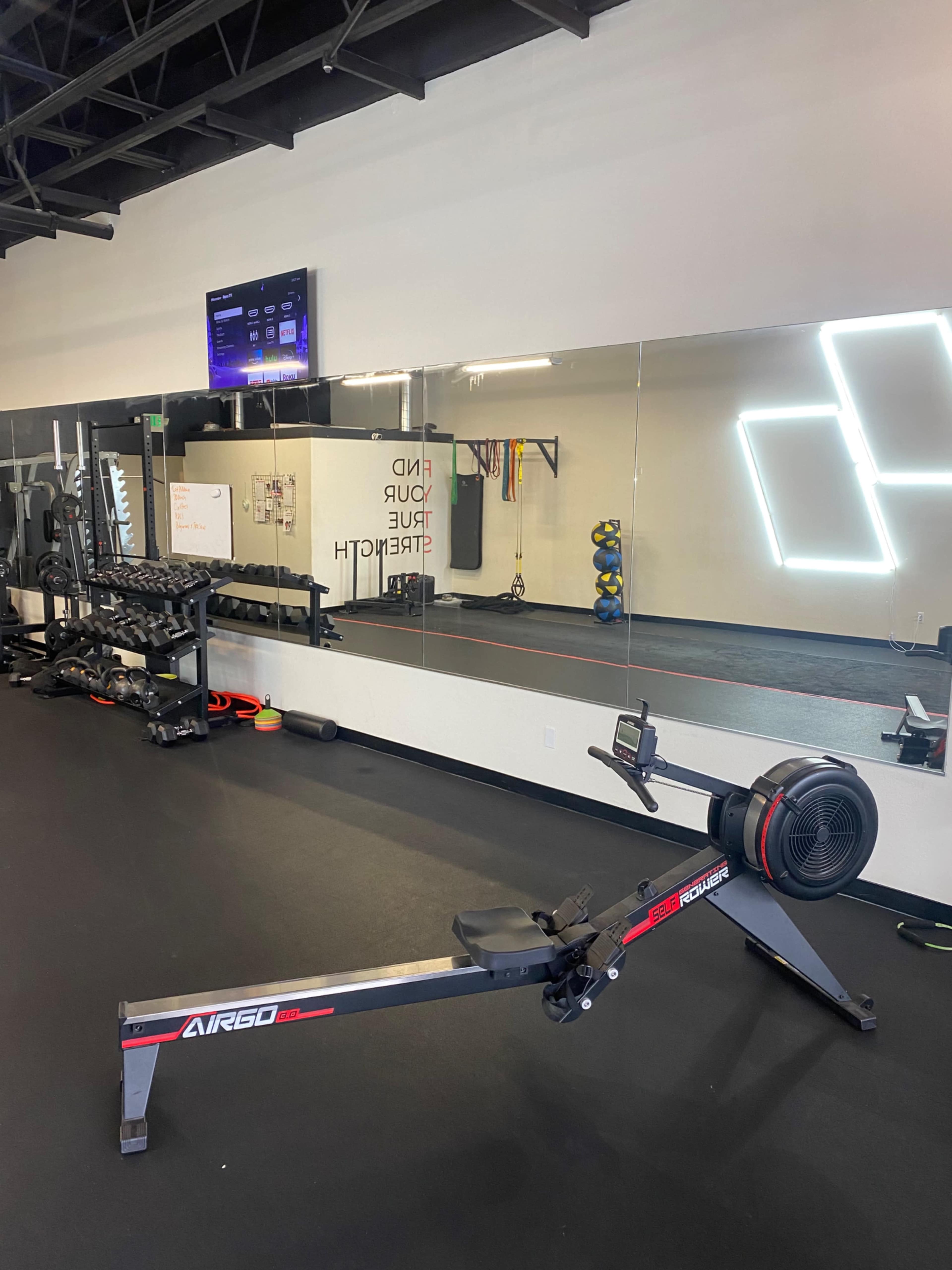 A gym interior with a rowing machine in the foreground, weightlifting equipment against the wall, and a mirror reflecting the space.