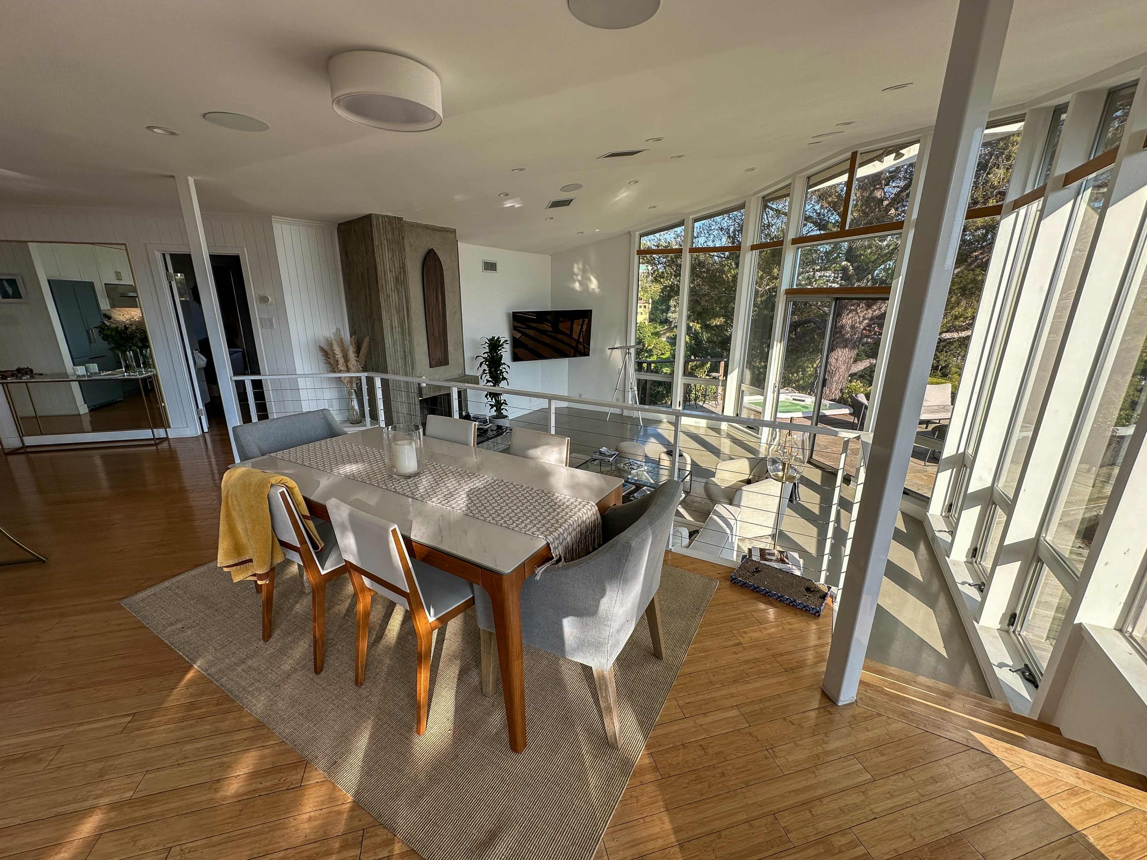 The image depicts a spacious, modern dining area with a large table surrounded by chairs, complemented by large windows that provide natural light and a view of the outdoors.