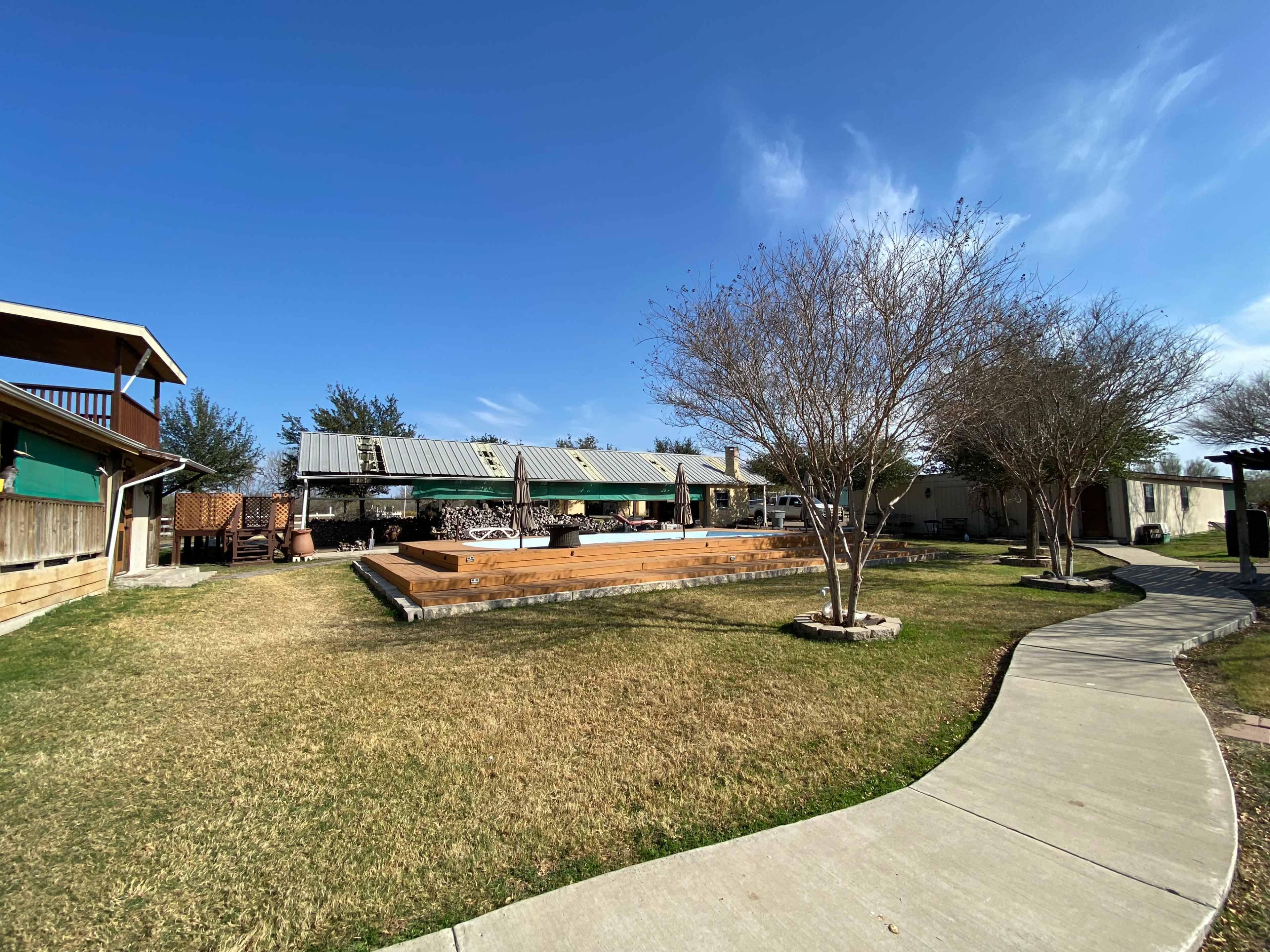 The image shows a landscaped outdoor area with a pathway leading to a wooden deck and several structures under a clear blue sky.