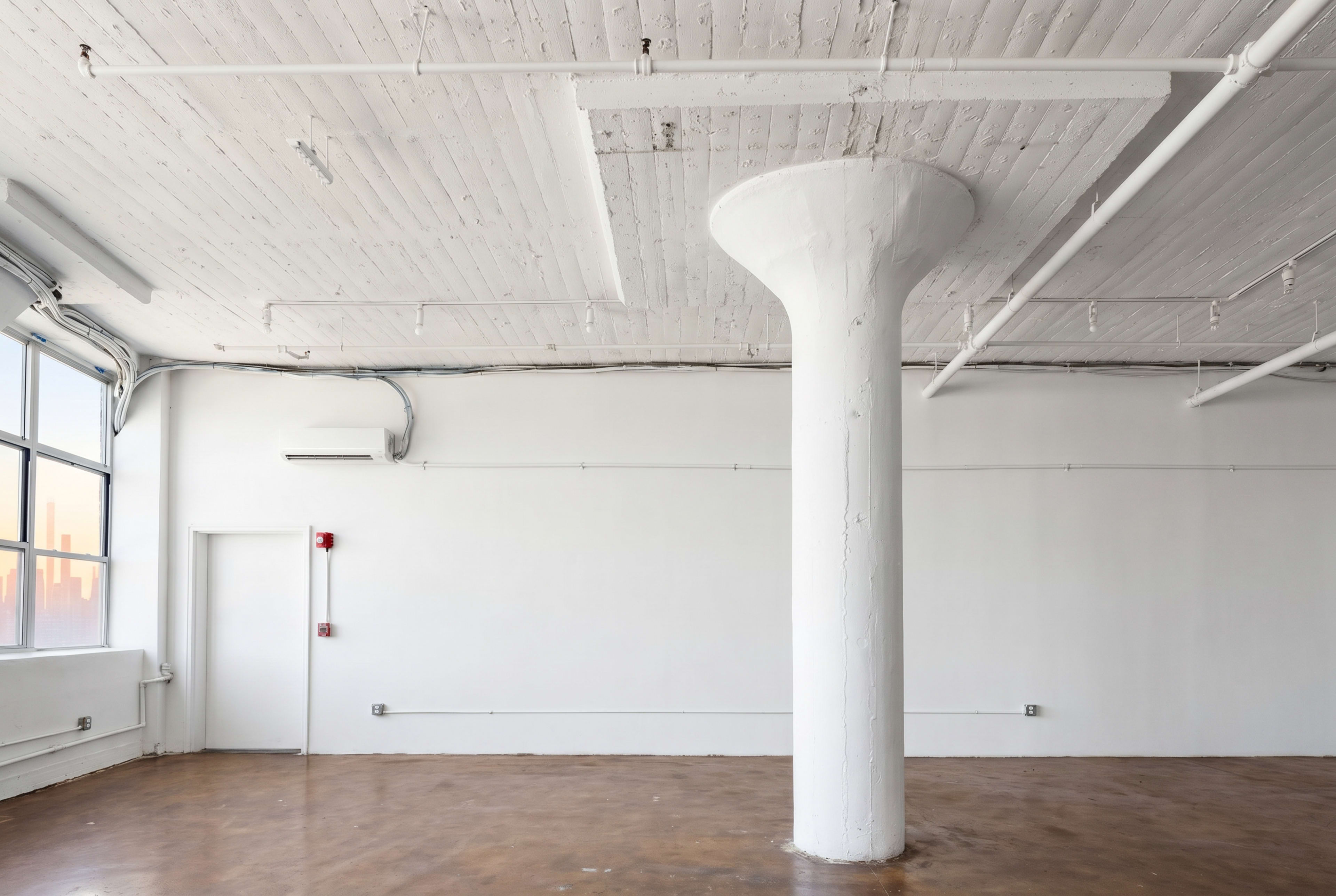 The image shows a spacious, empty room with a polished concrete floor and a central white column under a ceiling with exposed beams and pipes.