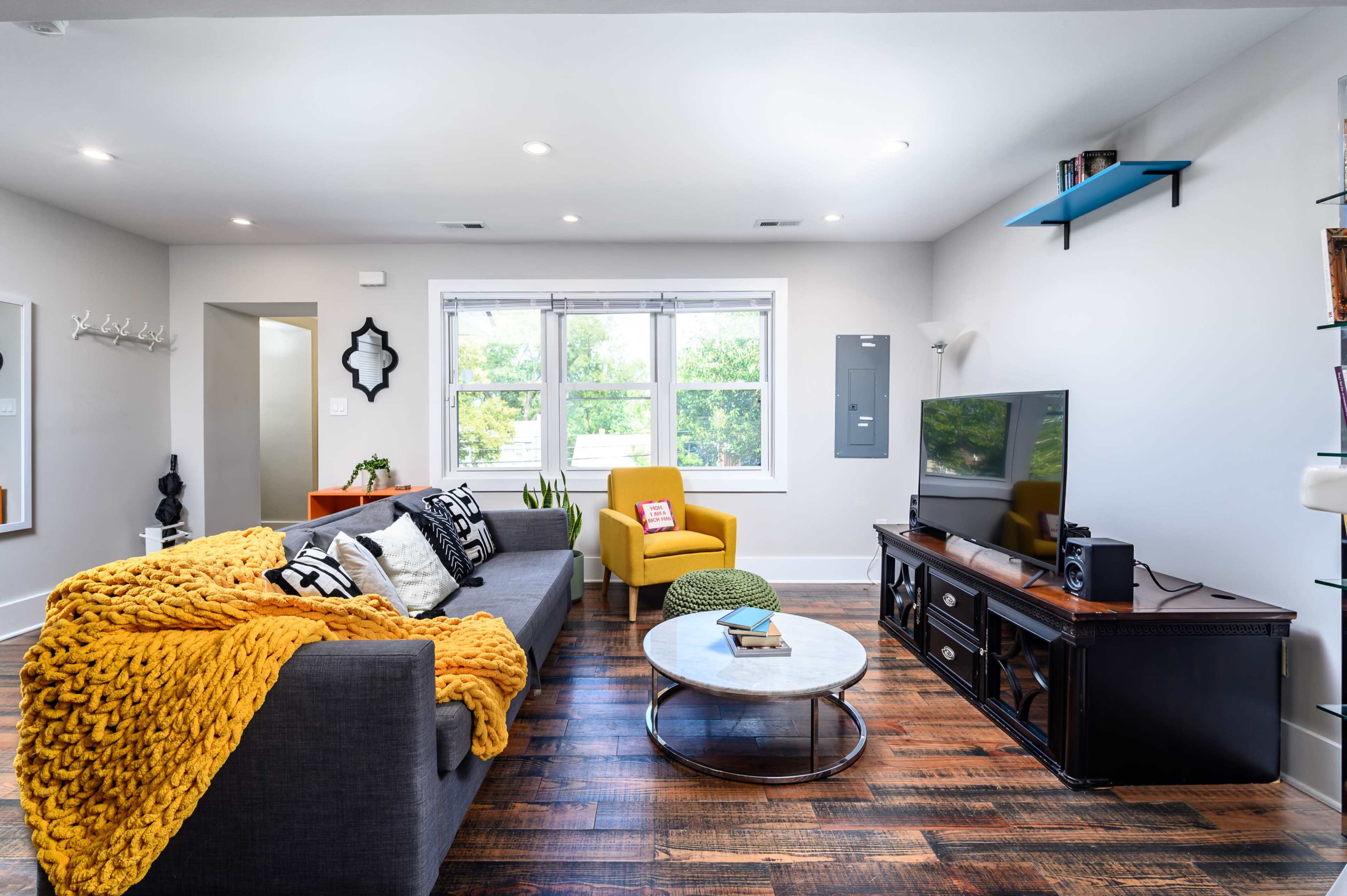 The living room features a gray sofa draped with a yellow blanket, a round coffee table, an armchair, and a television on a dark wood console, with large windows letting in natural light.