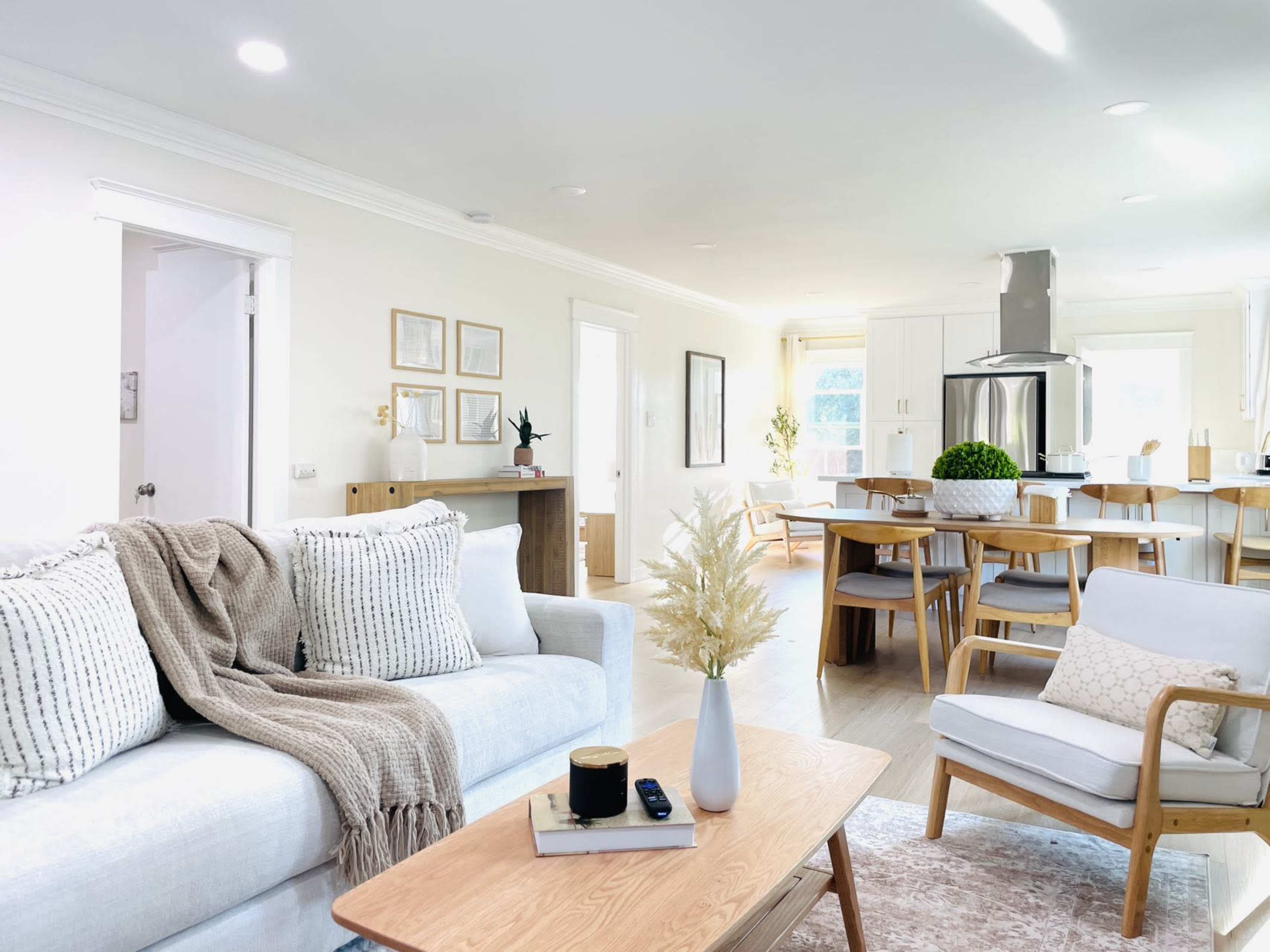 A modern living room features a light-colored sofa with decorative pillows, a wooden coffee table, and an open layout leading to a dining area with a large table and kitchen in the background.