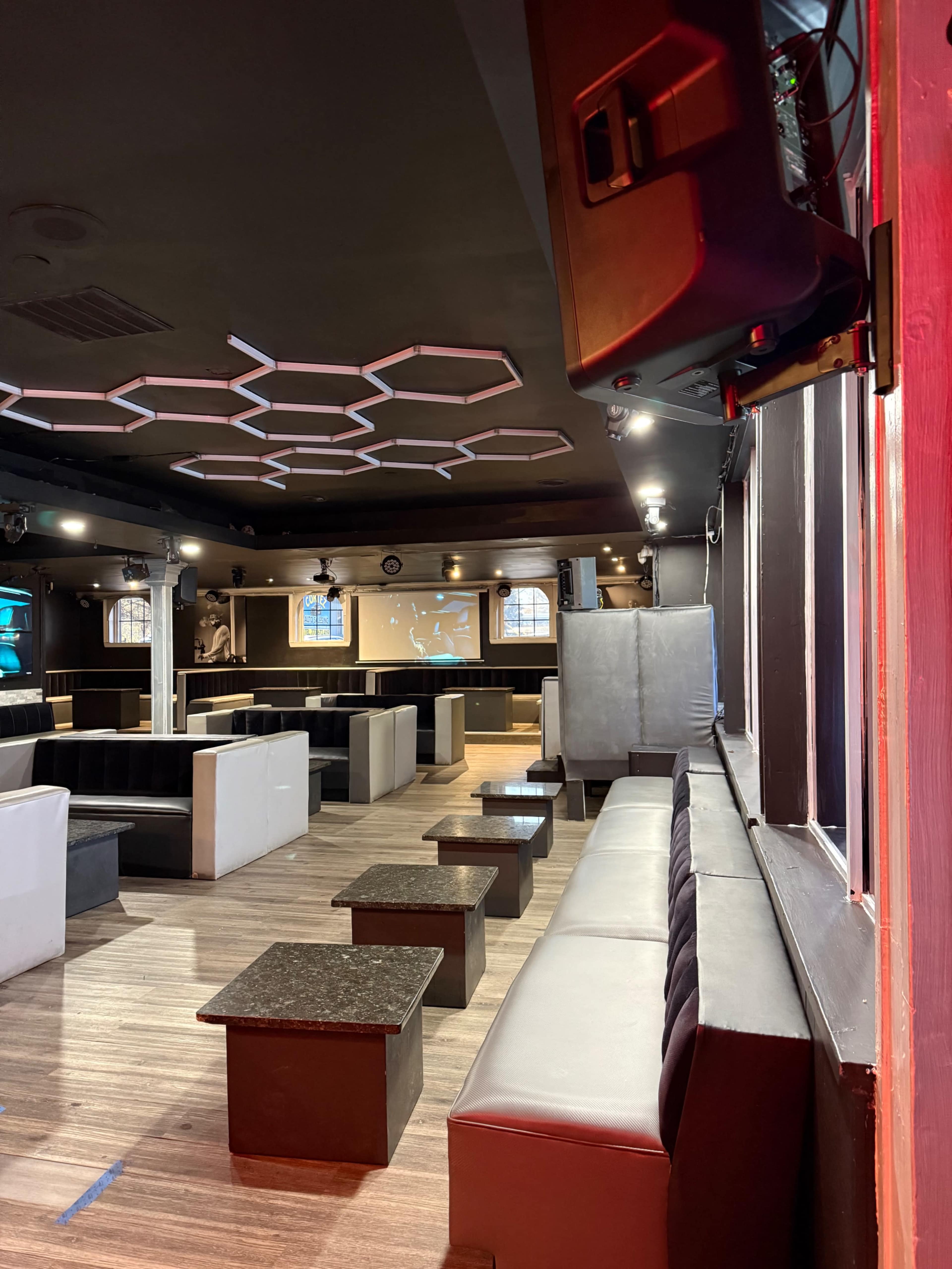 The image shows an empty lounge area with hexagonal ceiling lights, black and white seating arrangements, and small tables.