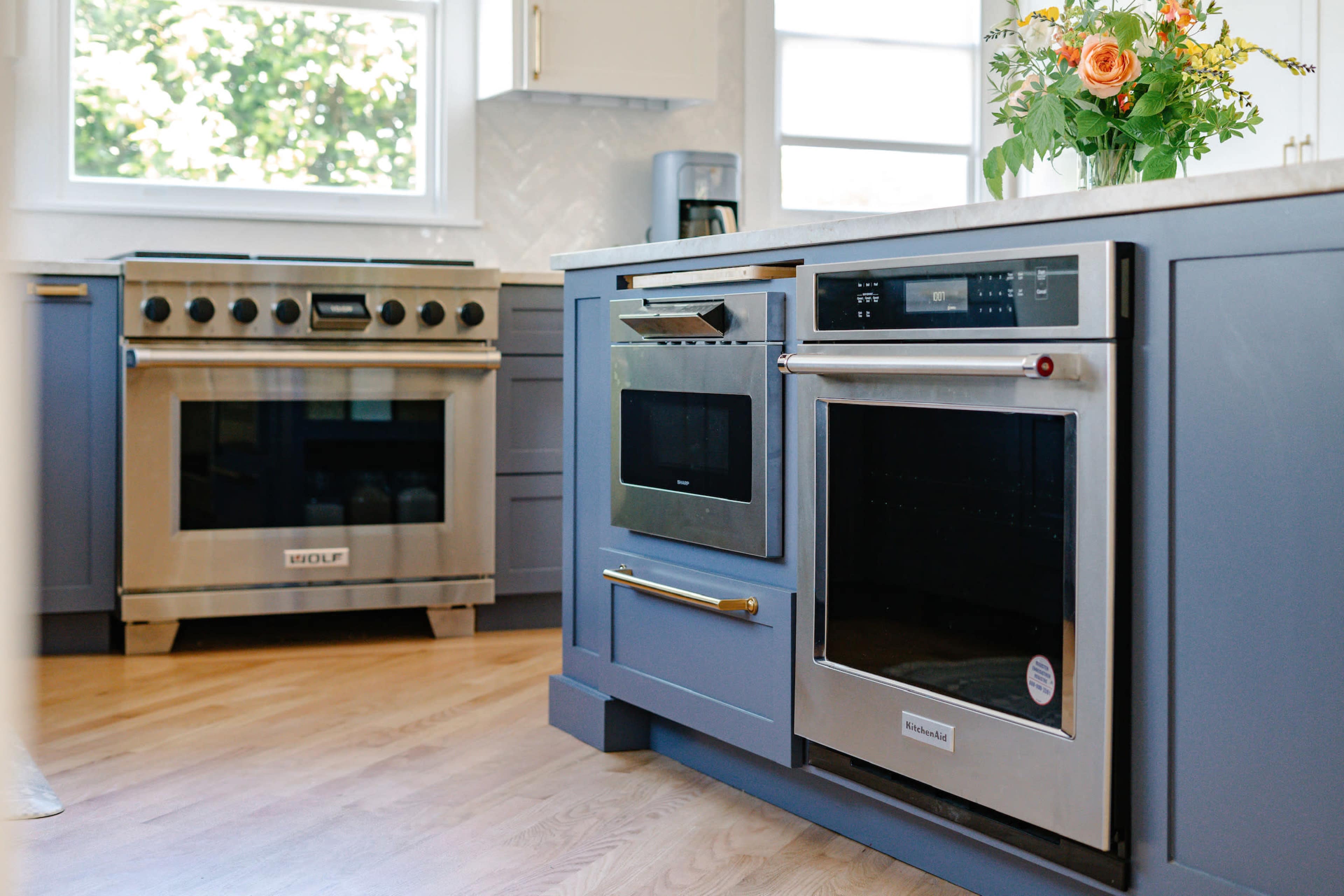 A modern kitchen features stainless steel appliances, including a range and an oven, with blue cabinetry and a window providing natural light.