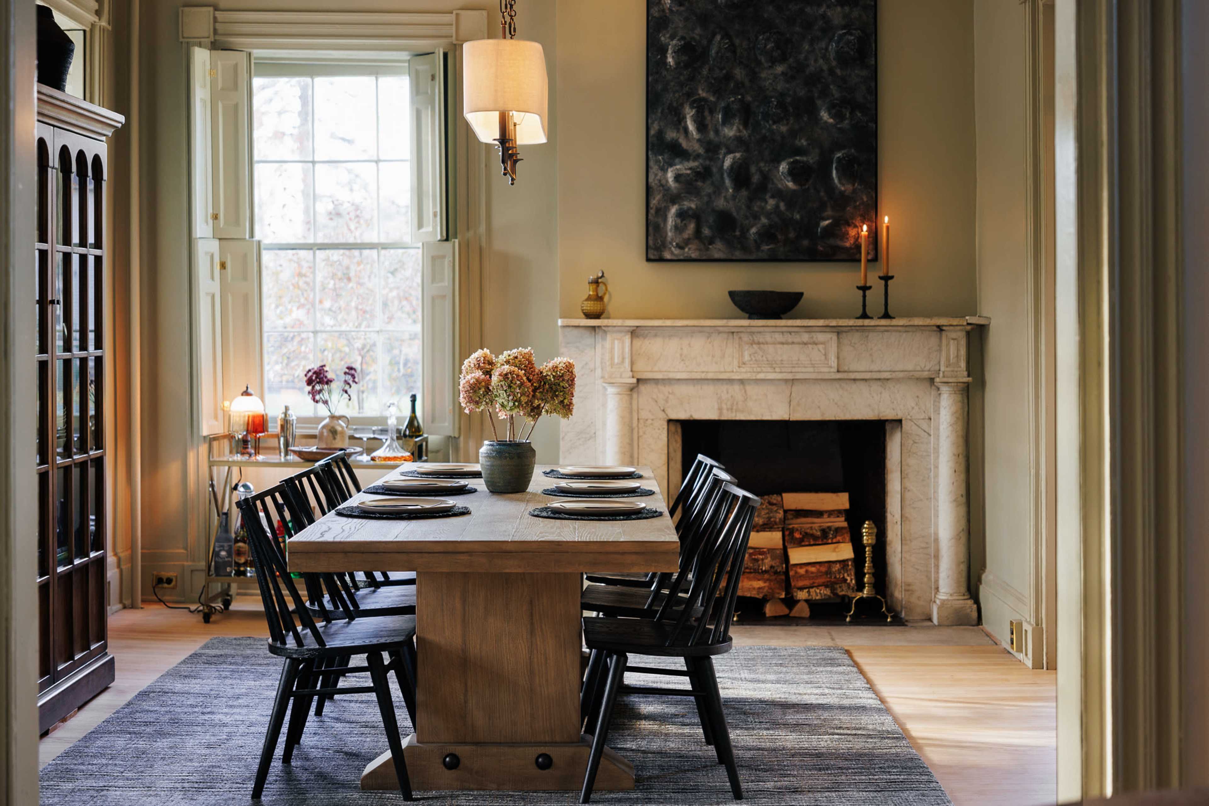 A dining room features a wooden table surrounded by black chairs, with a fireplace and a large artwork on the wall.