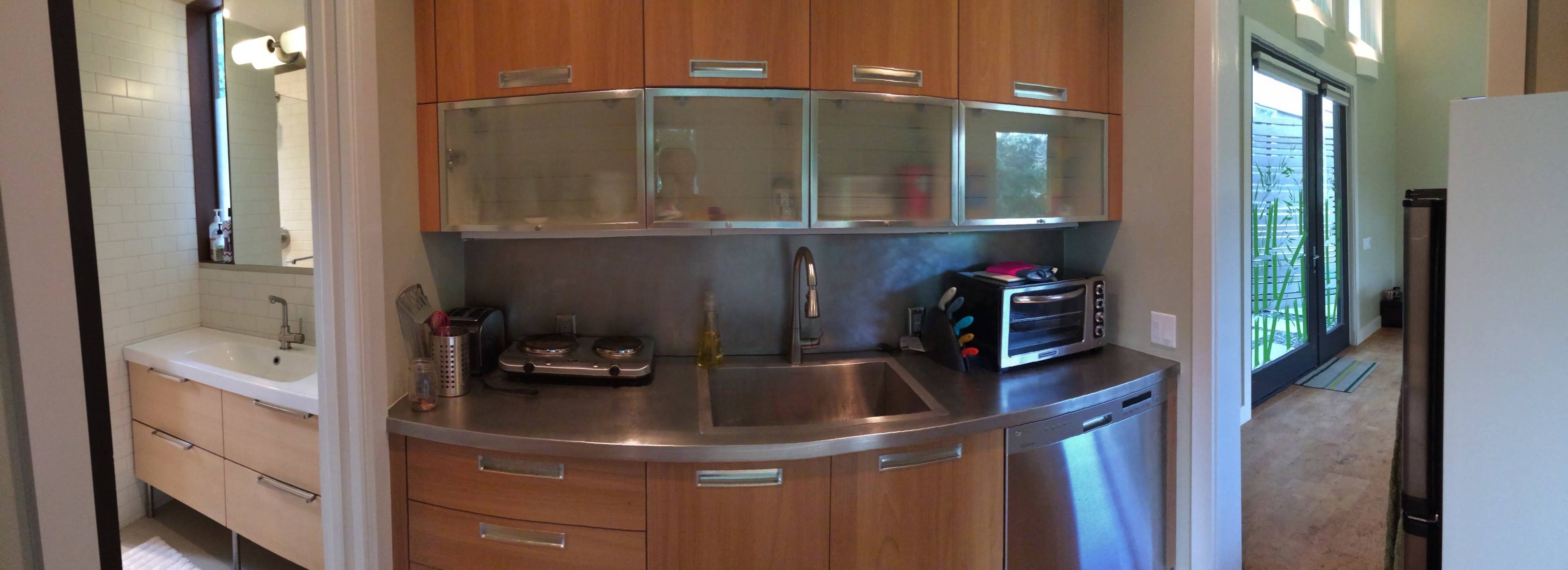 A modern kitchen with stainless steel countertops and wooden cabinetry, adjacent to a bathroom visible on the left and an outdoor space accessible through a sliding door on the right.