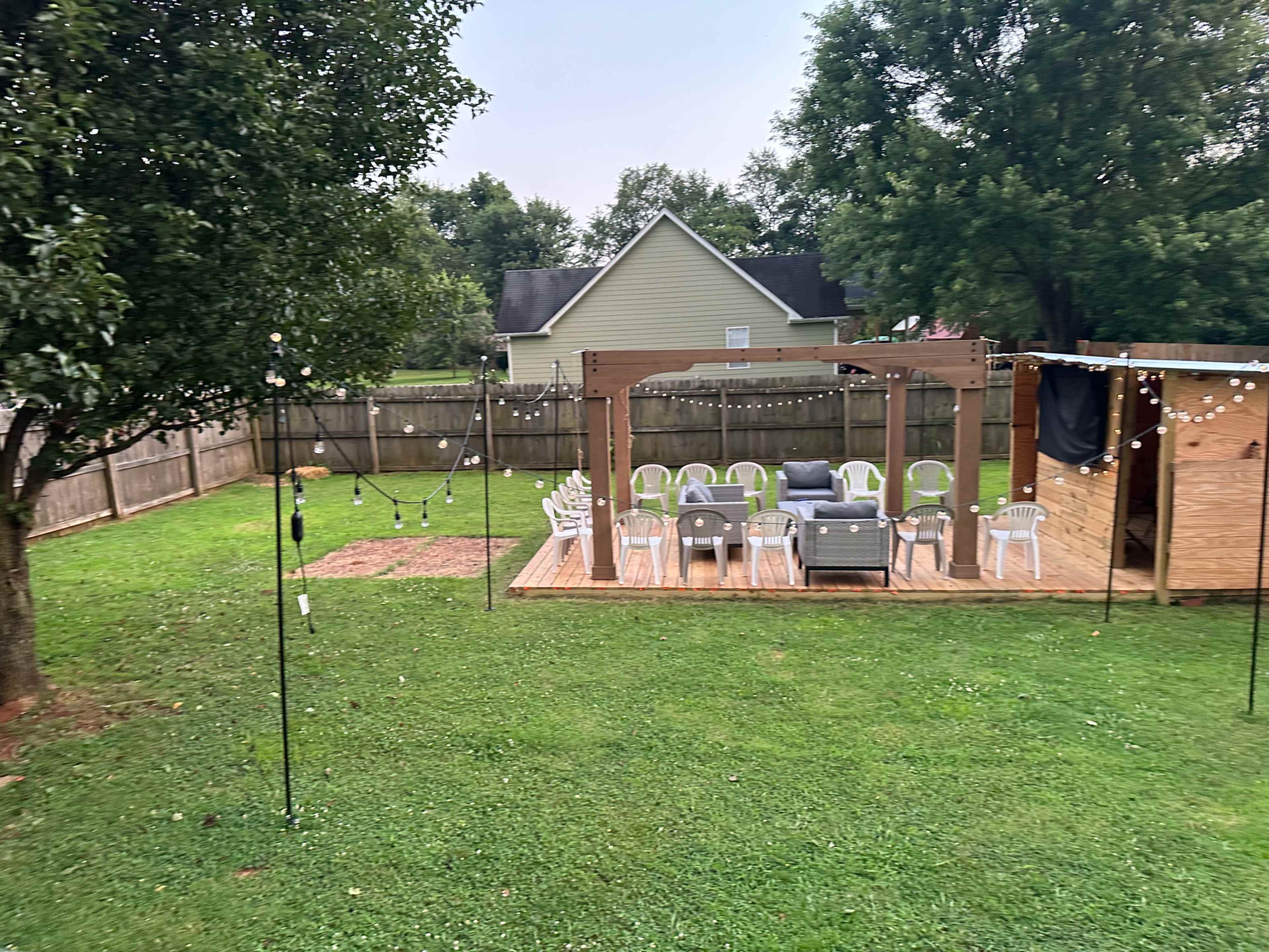 A grassy backyard with a wooden deck containing chairs and a couch, surrounded by a fence and trees.