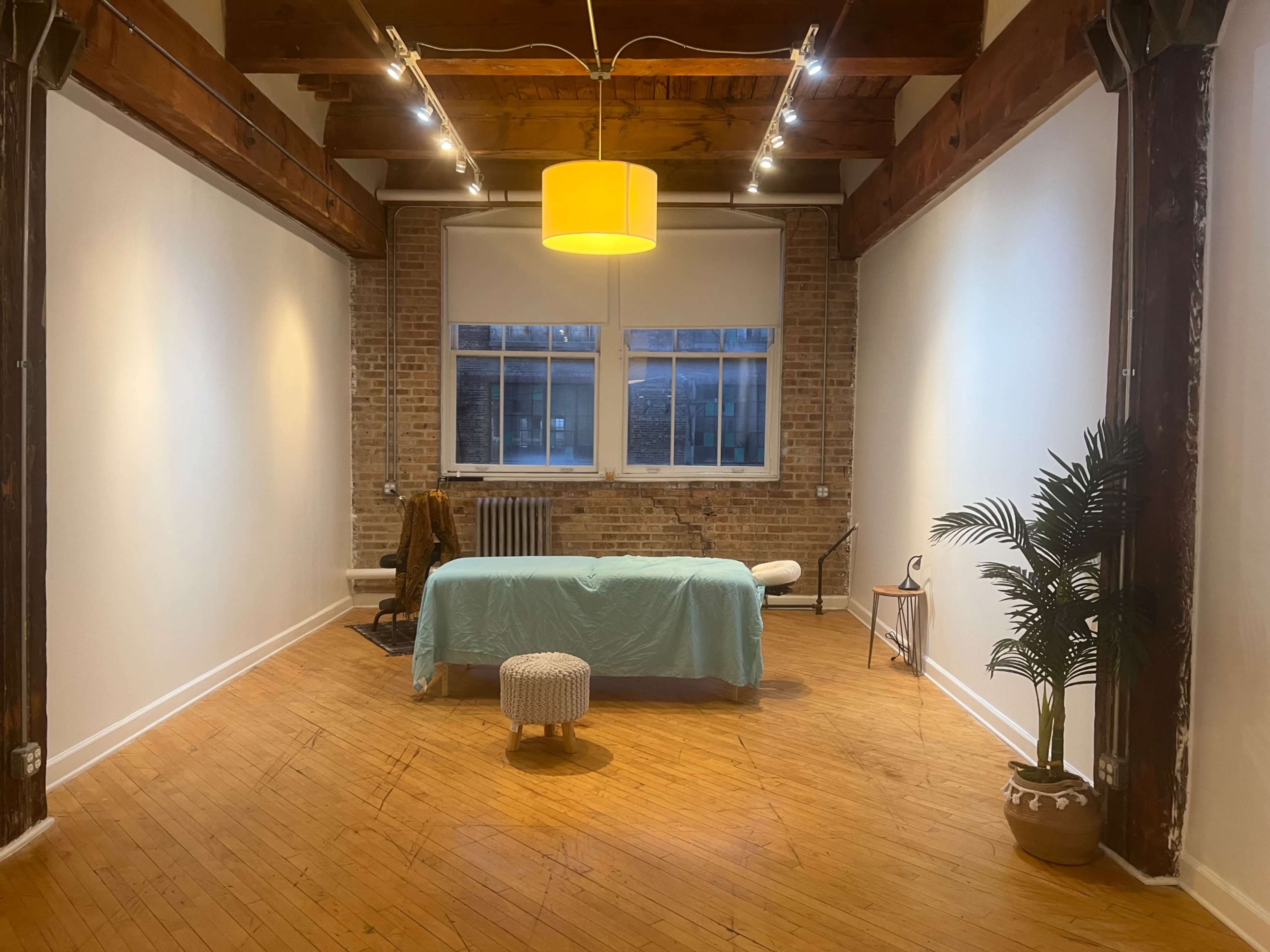 The image shows a spacious, well-lit room with wooden floors, featuring a massage table covered with a light blue sheet, a small stool, a potted plant, and large windows on the brick wall.