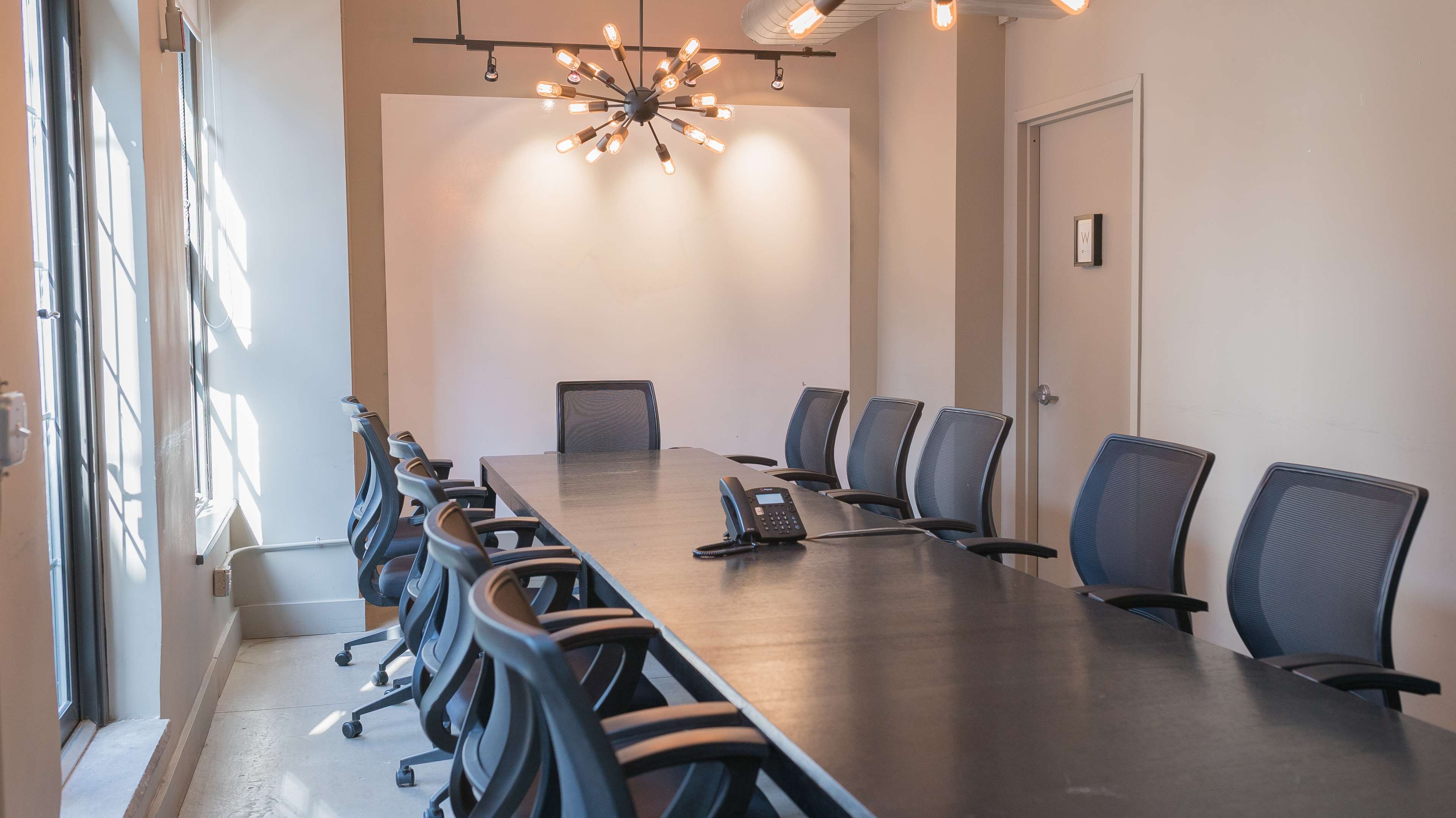 A large conference room features a long table surrounded by ergonomic chairs and a central light fixture.