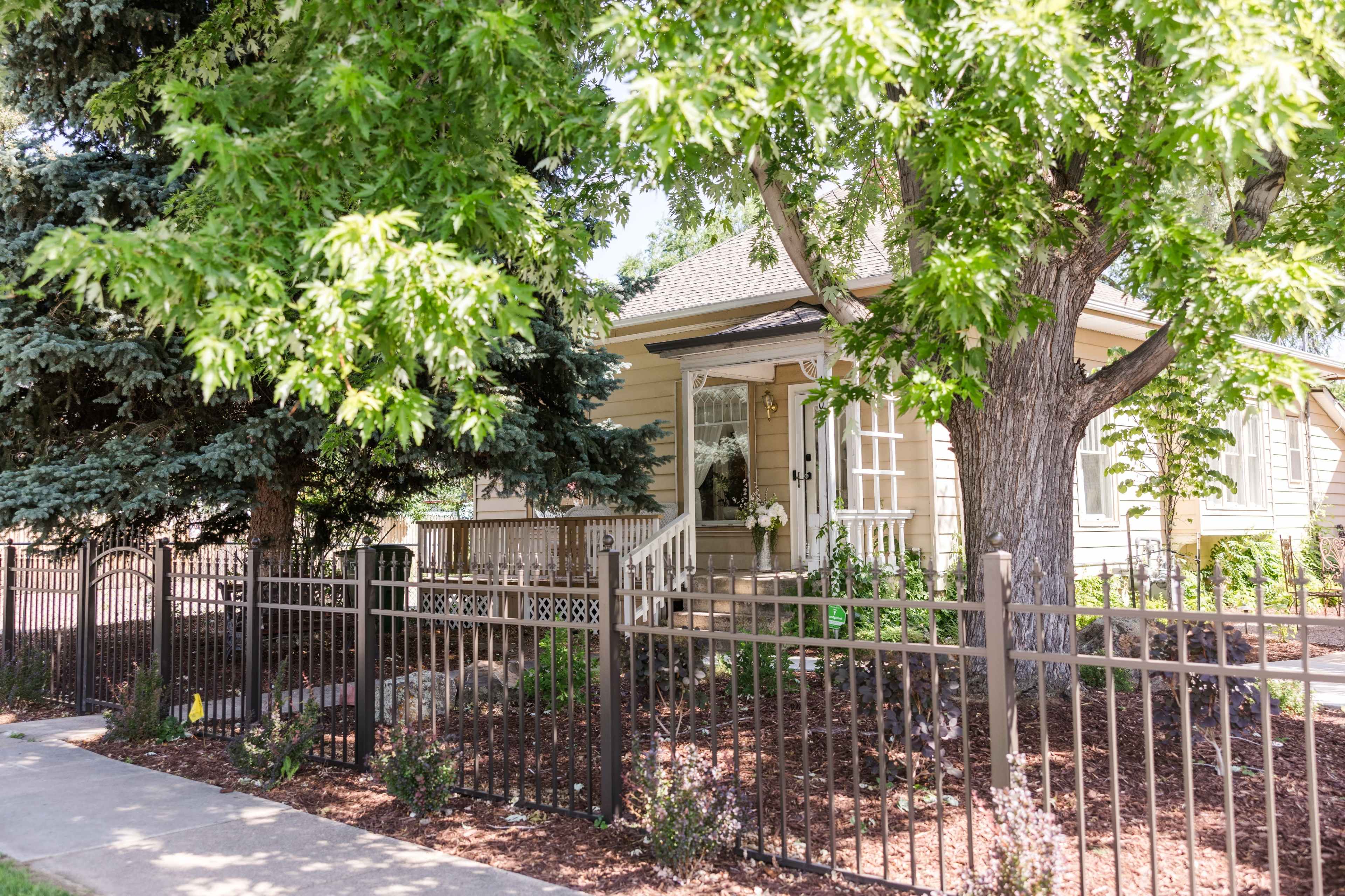 A small, light-colored house with a porch is surrounded by greenery and a black metal fence.