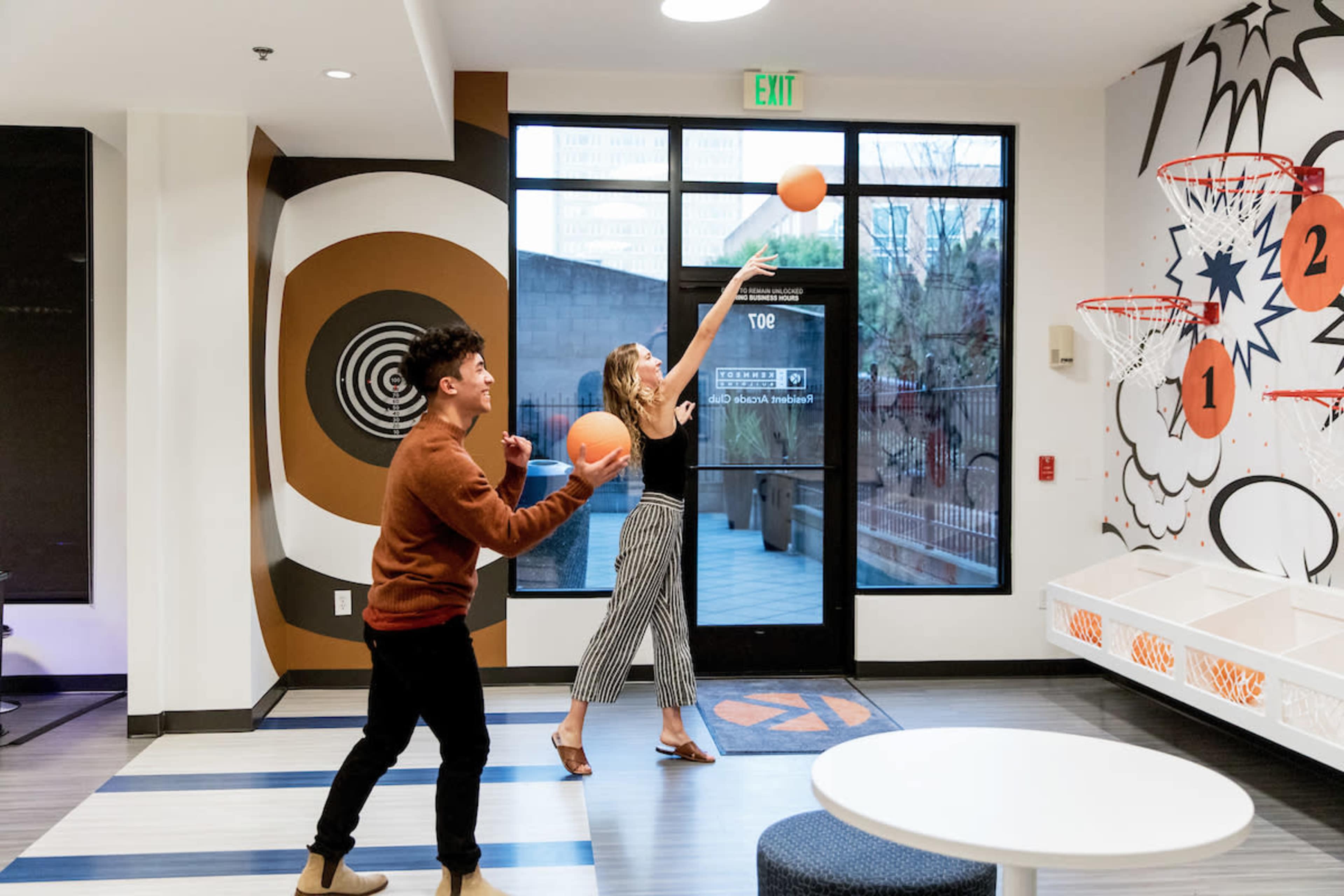 Two people are playing basketball in a recreational room with a large window and decorative wall graphics.