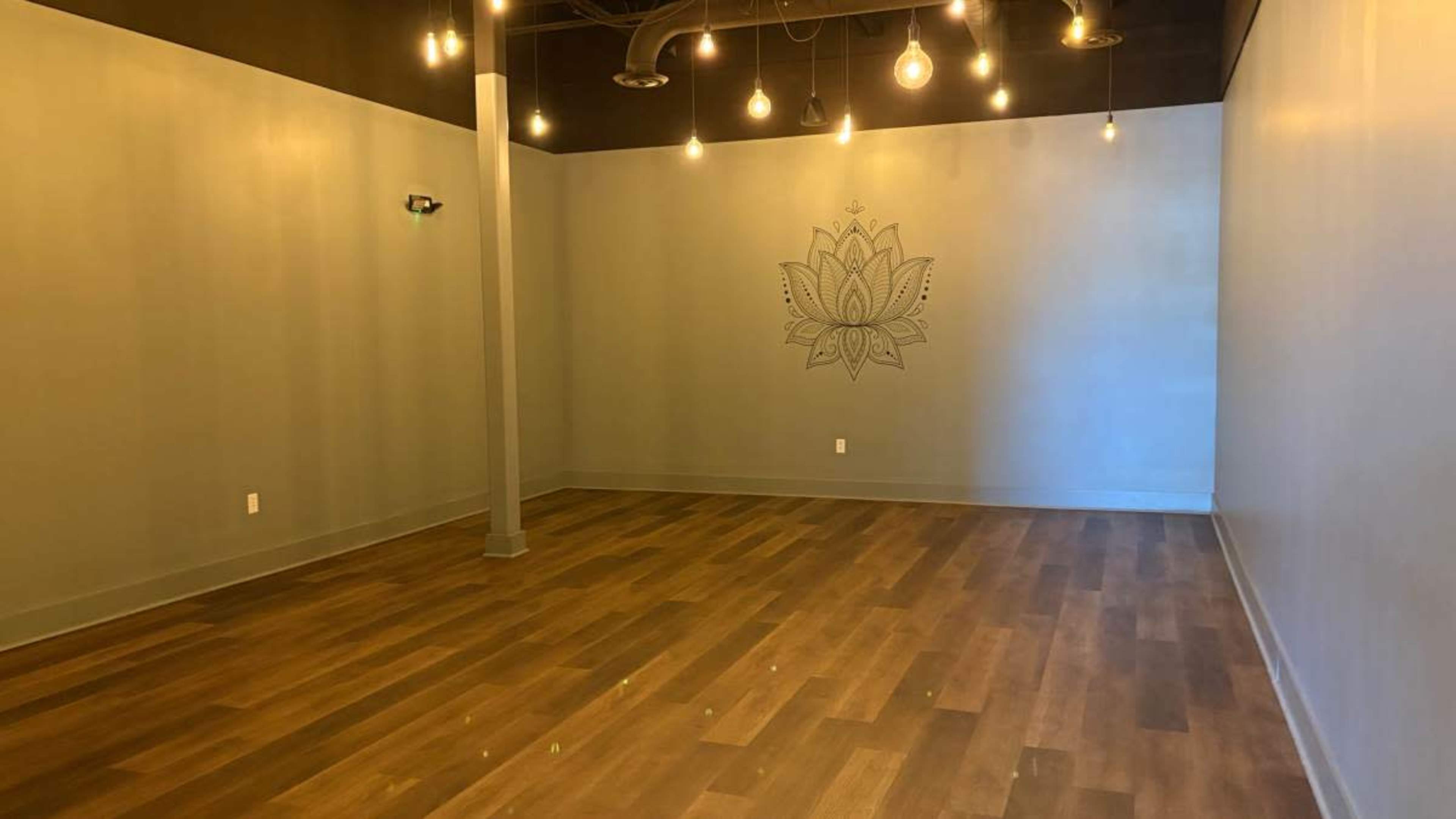 The image shows a spacious, empty room with wooden floors, a lotus design on the wall, and pendant light fixtures hanging from the ceiling.