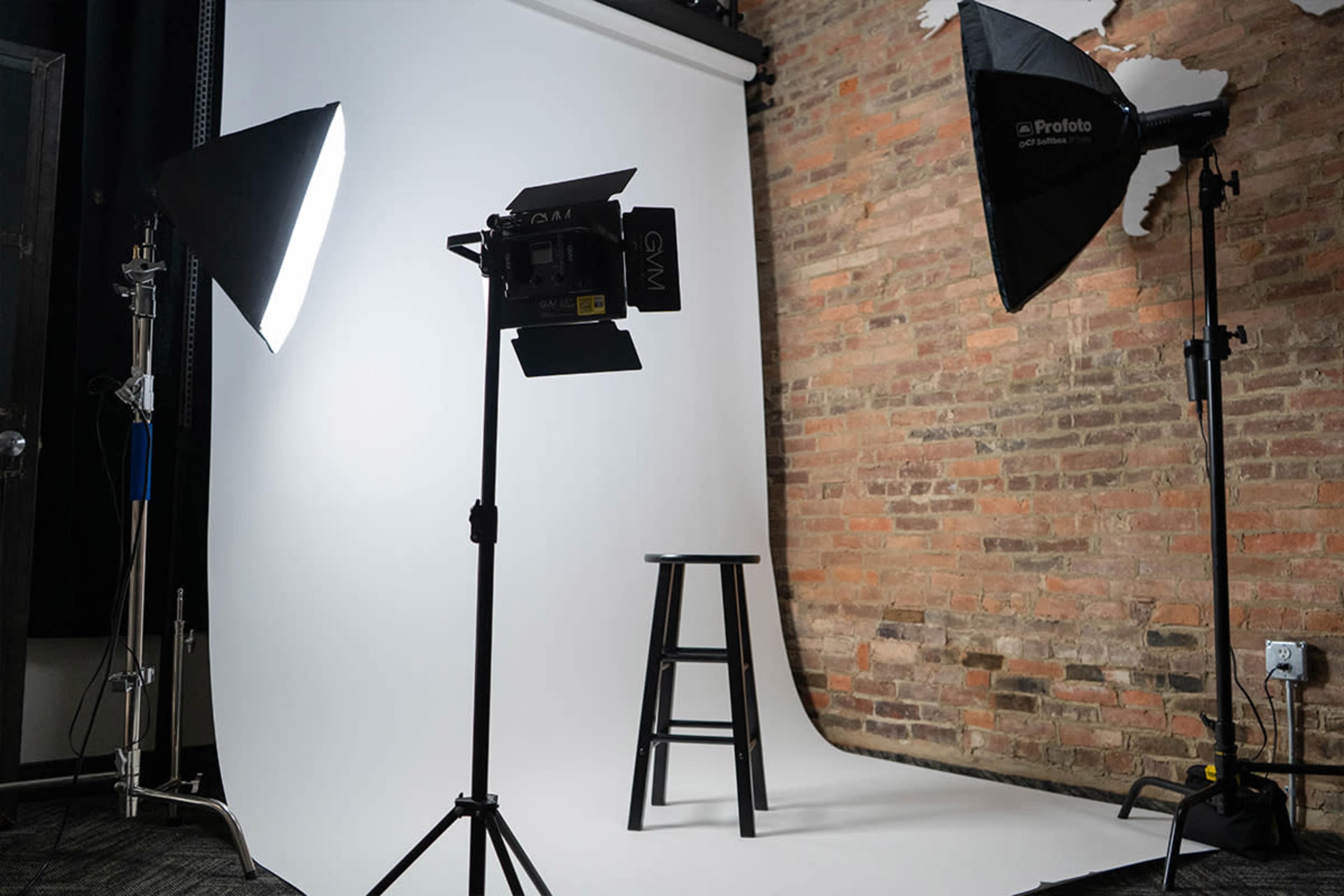 The image shows a photography studio setup featuring a white backdrop, two softbox lights, and a black stool positioned in the center.