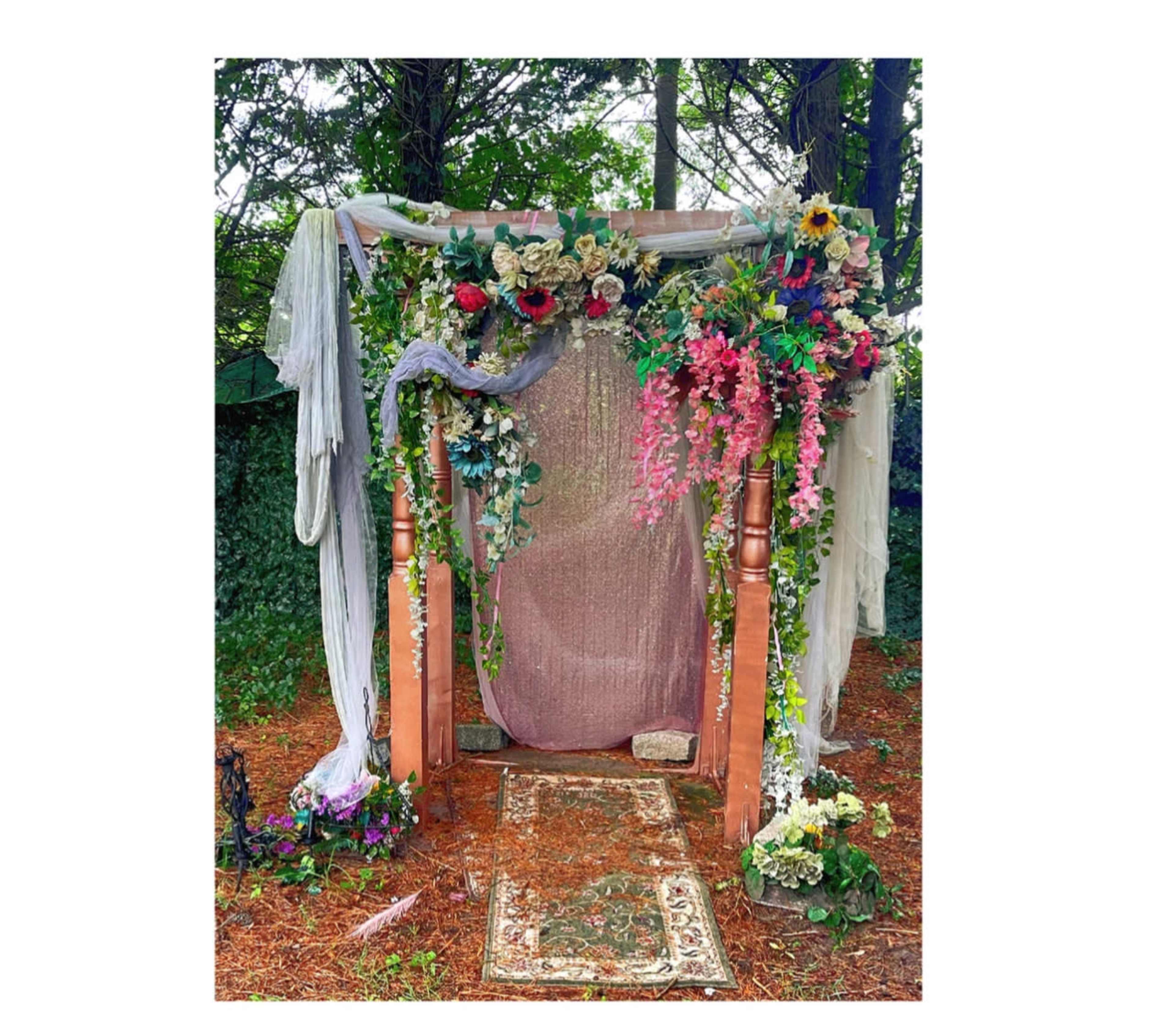 A floral-decorated archway with a draped fabric backdrop and a patterned rug on the ground in a natural setting.