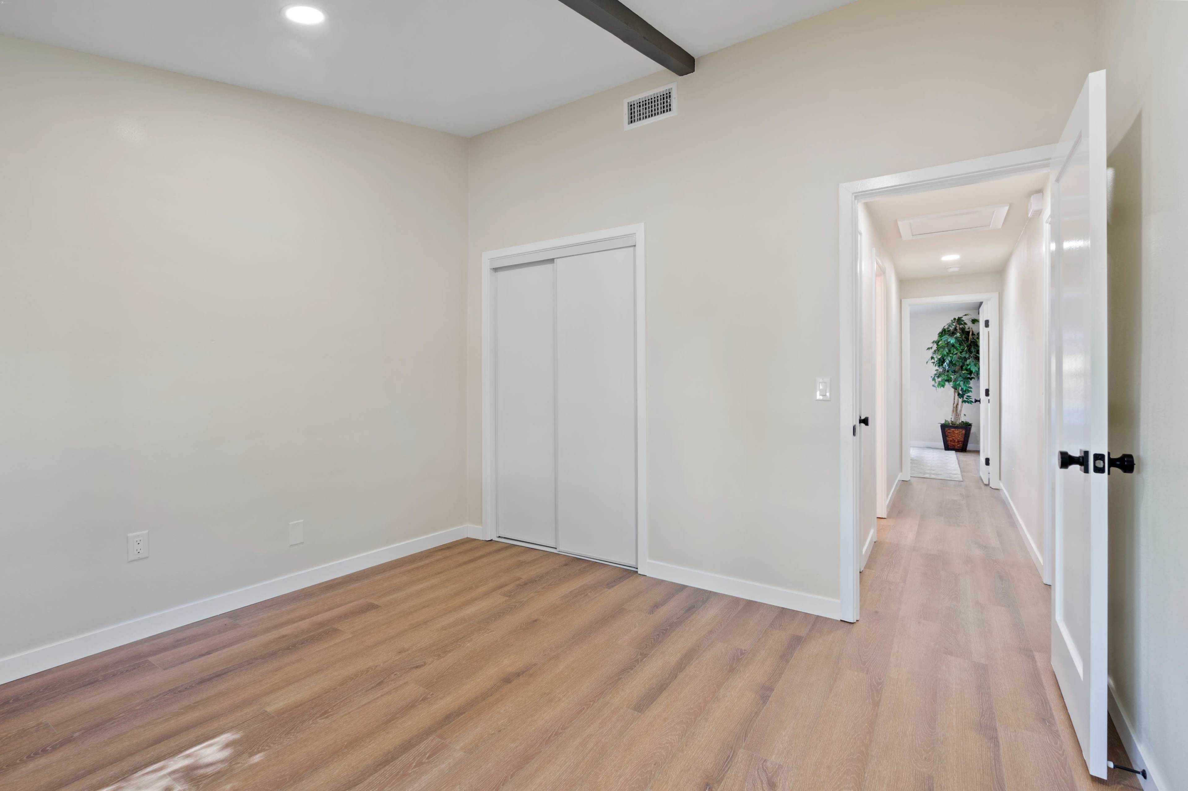 The image shows a spacious, empty room with light-colored walls and a large closet, leading to a hallway with potted plants.