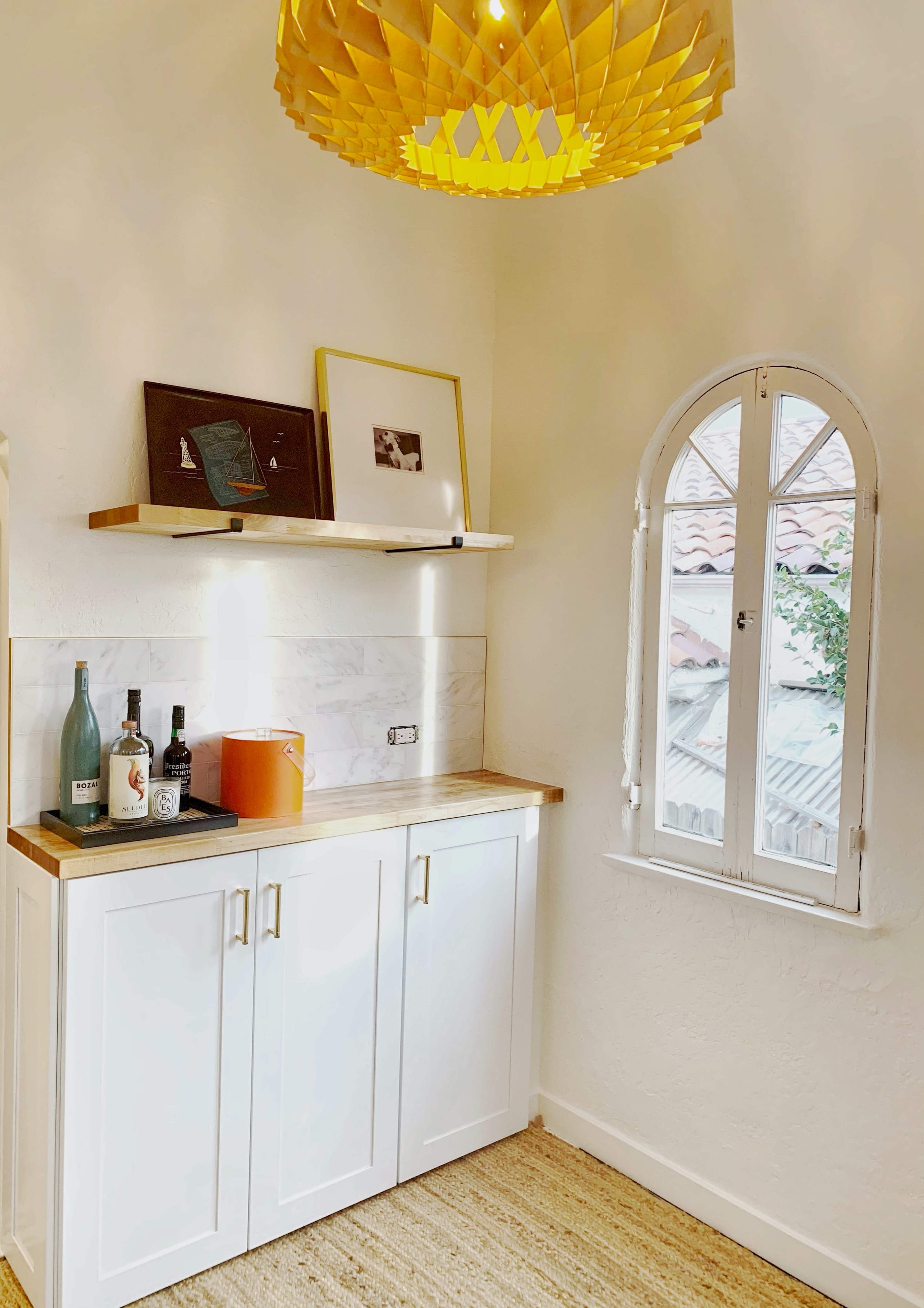 A well-lit corner of a room features white cabinetry, a wooden countertop, a decorative orange container, bottles of liquor, and a large, unique pendant light above.