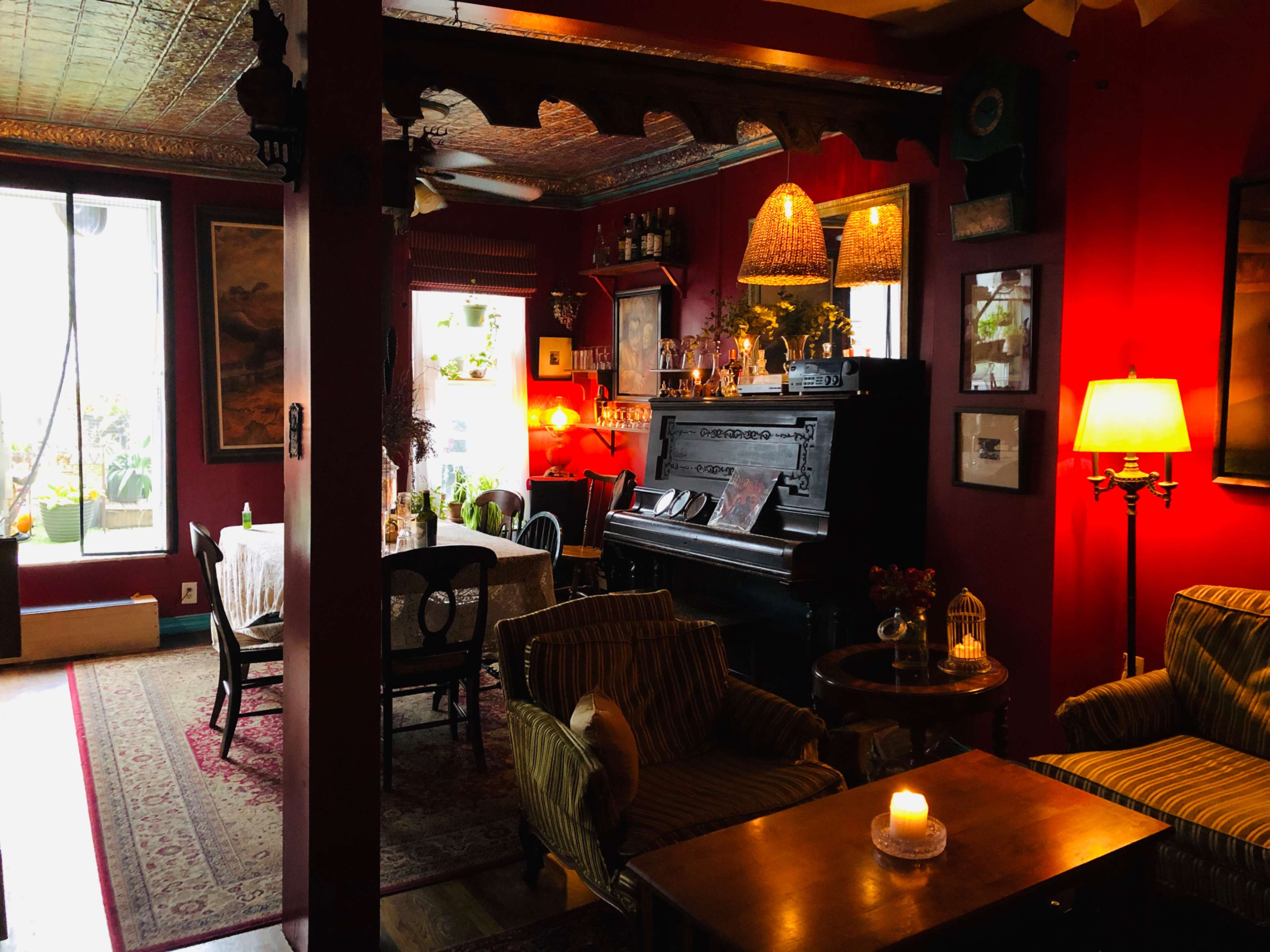 The image shows a warmly lit, cozy lounge area featuring a piano, comfortable seating, and decorative plants against a backdrop of red walls.
