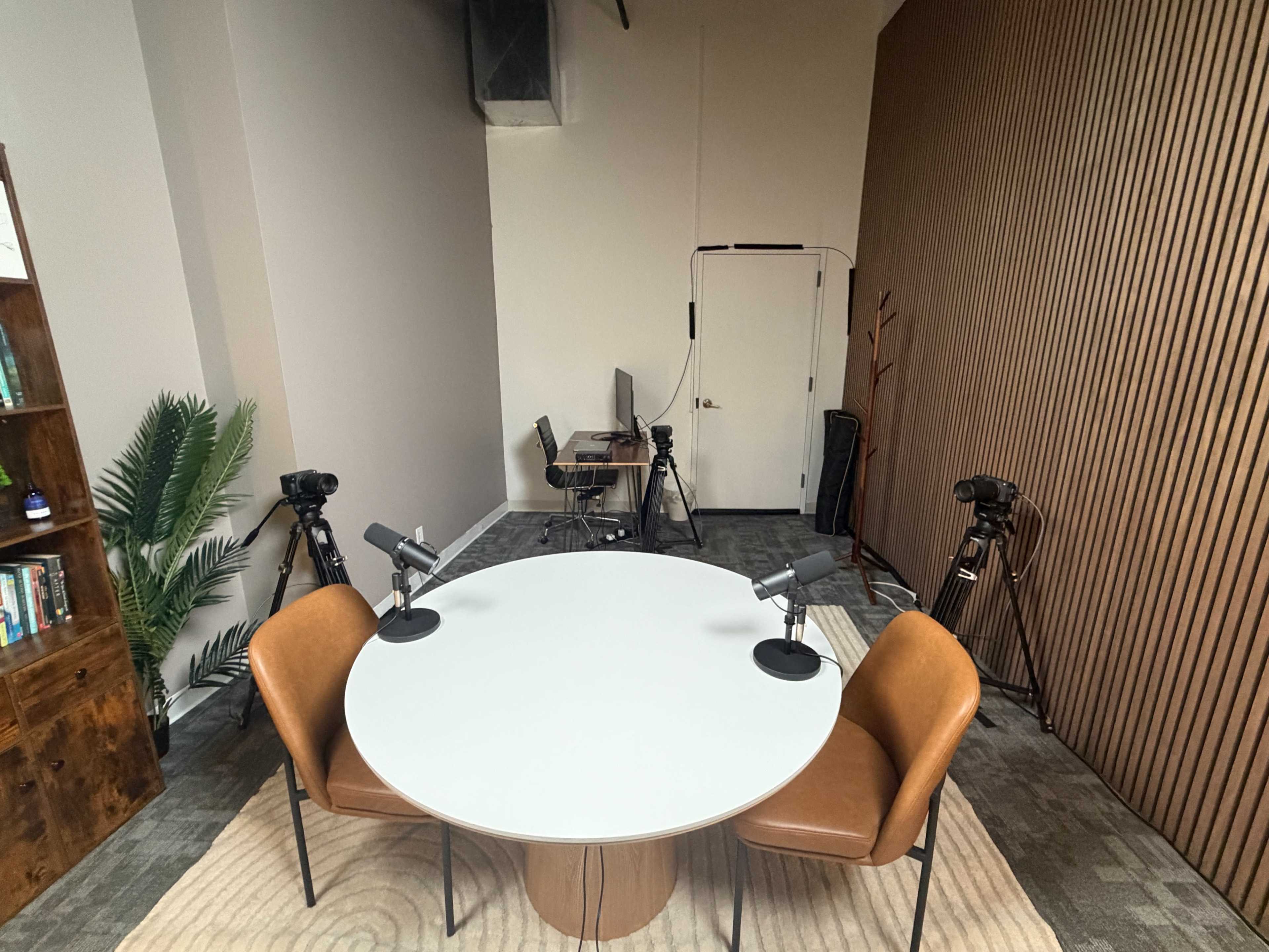 The image shows a small studio setup featuring a round table with two microphones, surrounded by chairs, and several cameras positioned around the room.