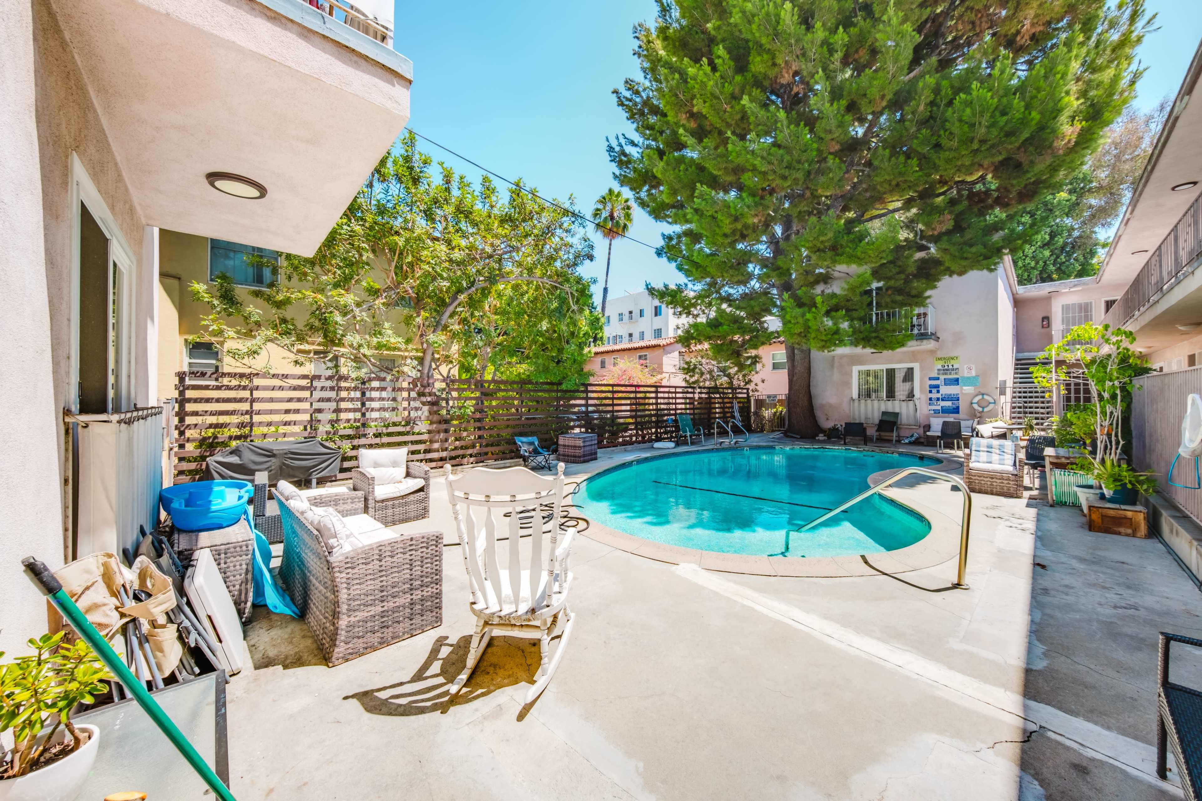 The image shows a swimming pool surrounded by a patio area with lounge chairs and potted plants, situated next to a building and trees.