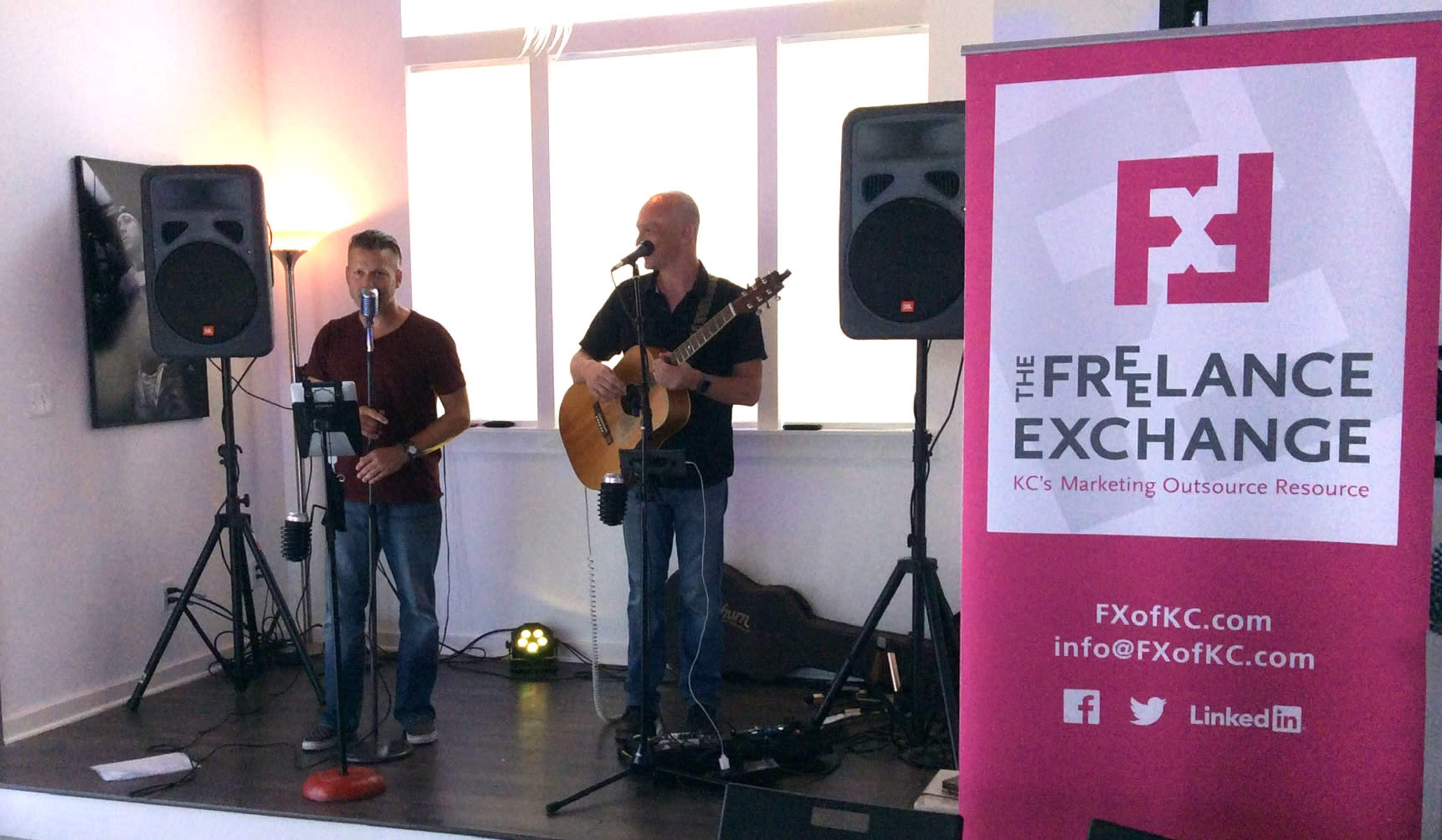 Two musicians are performing with a guitar and microphone on a small stage beside a banner for The Freelance Exchange.